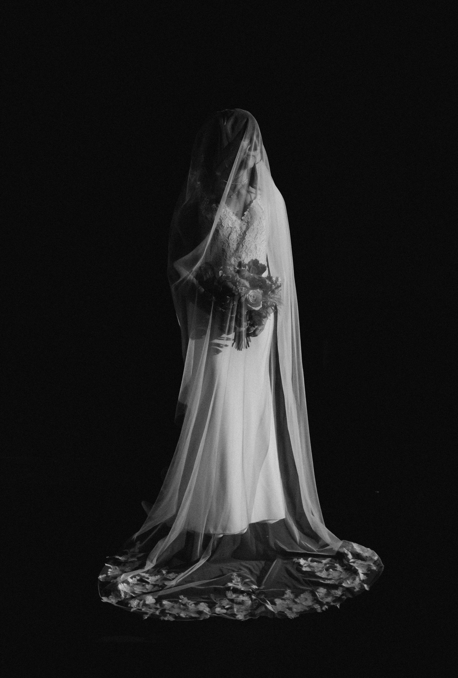 Black and white photo of a bride in a flowing gown with a long floral-trimmed veil holding a bouquet against dark background.