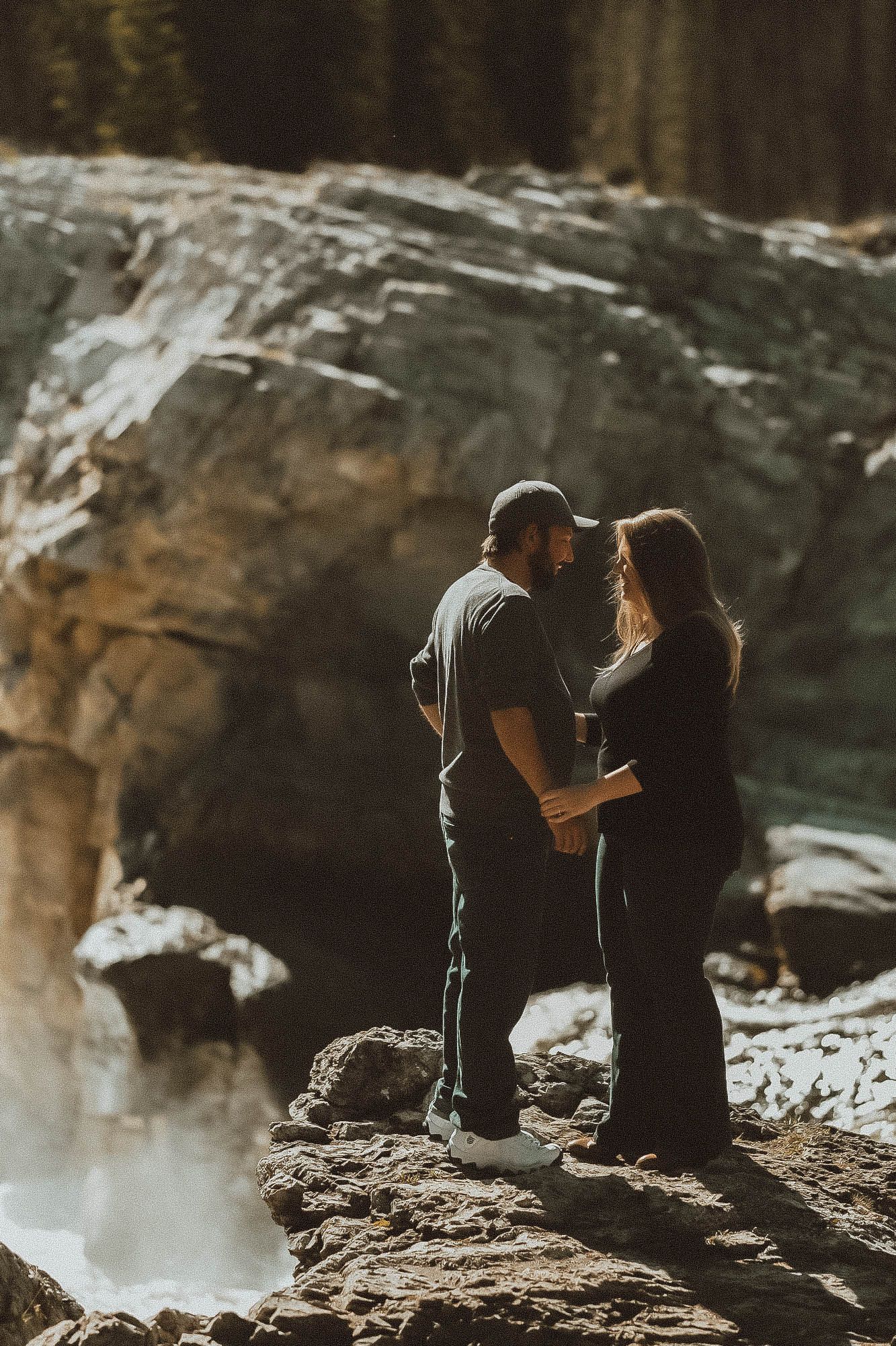 Couple sharing an intimate moment on rocky terrain near a waterfall, surrounded by dramatic mountain scenery.