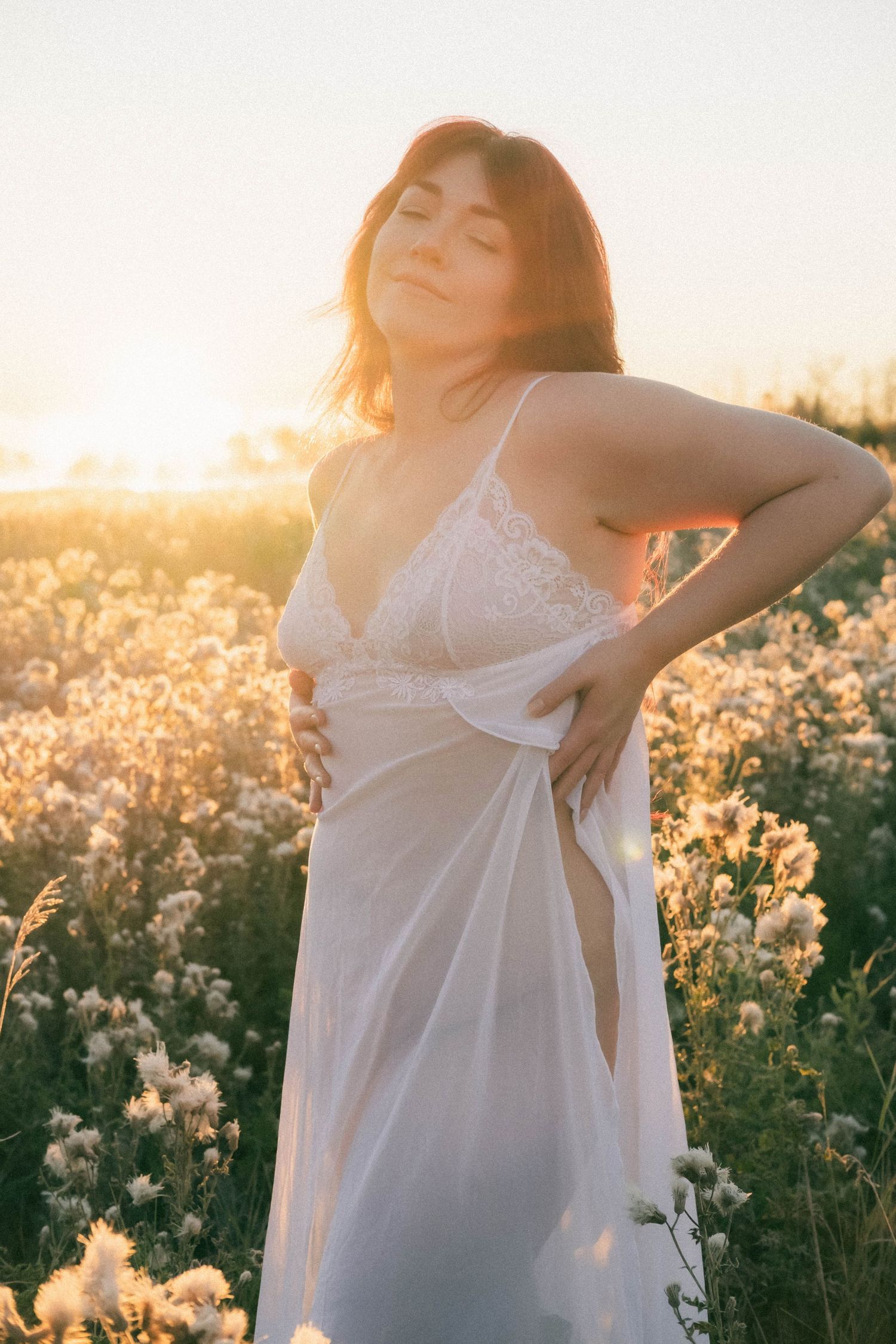 Woman in white lace dress standing in a sunlit flower field at golden hour with eyes closed.
