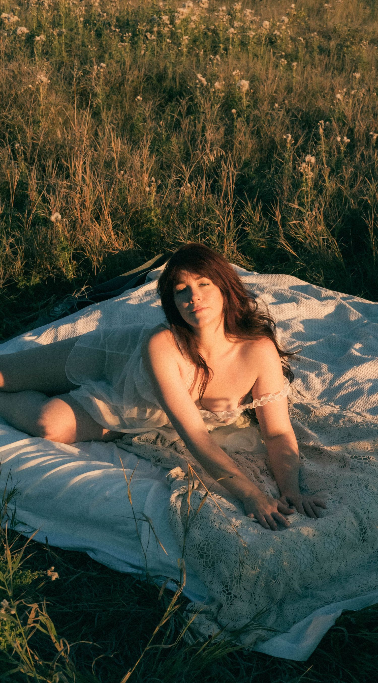 A woman with dark hair reclines on a white blanket in a sunlit wildflower field wearing a delicate white dress.