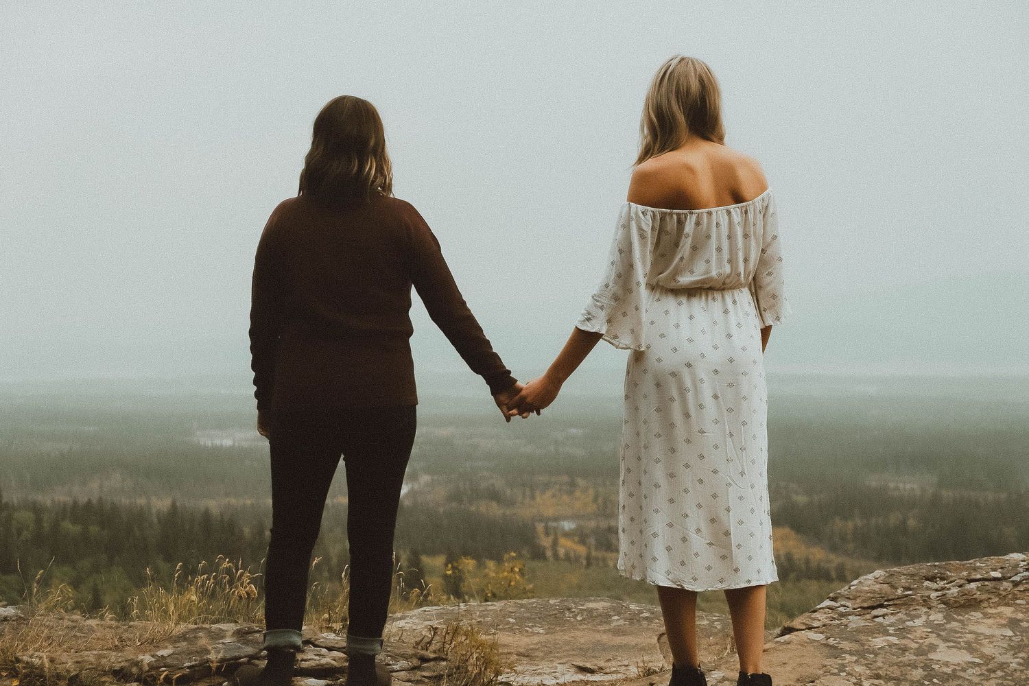 Two women holding hands while standing on a rocky overlook, gazing at a vast misty landscape of forests and hills.