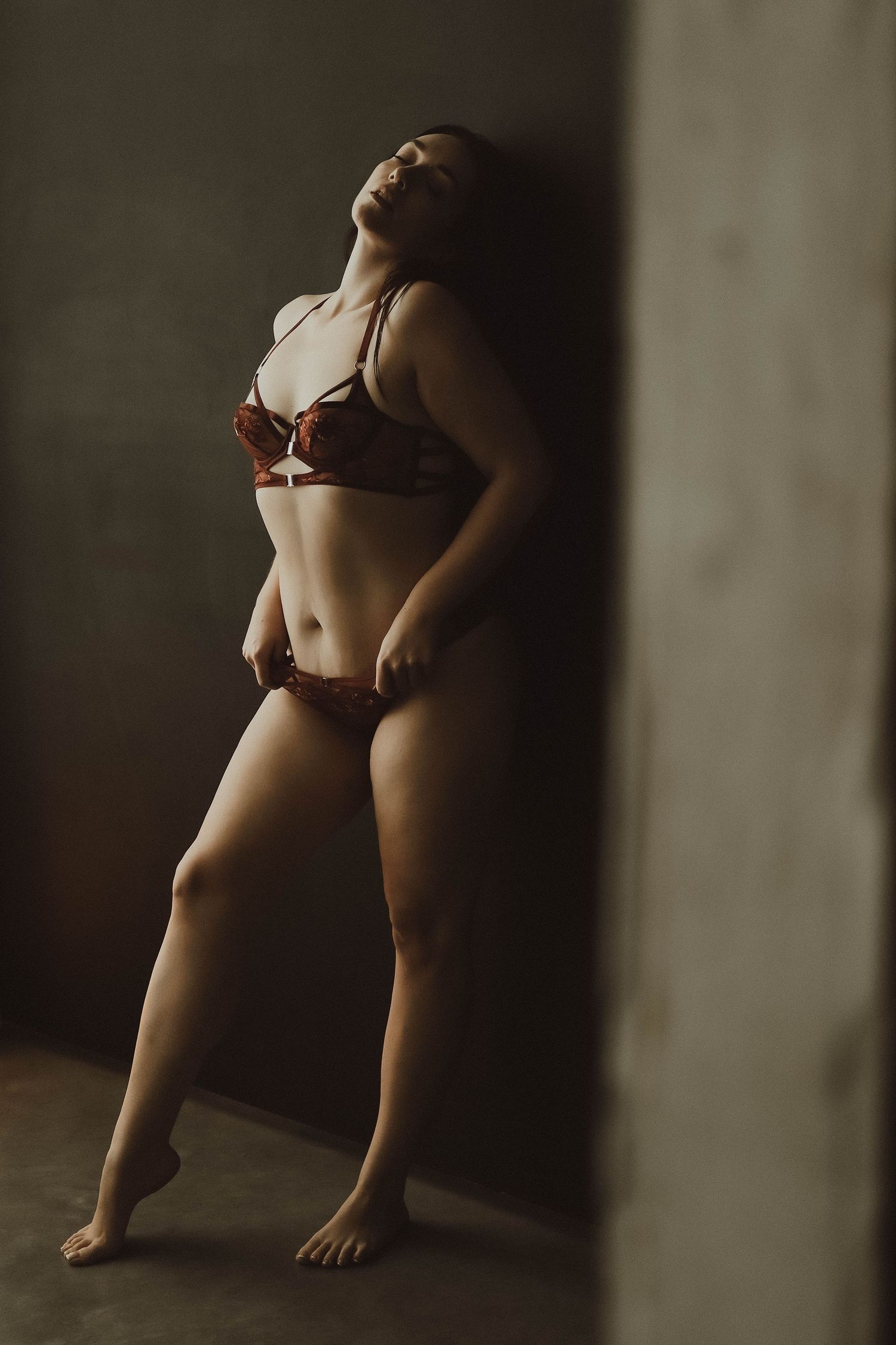 Woman in dark red lingerie posing confidently against a concrete wall in moody studio lighting.