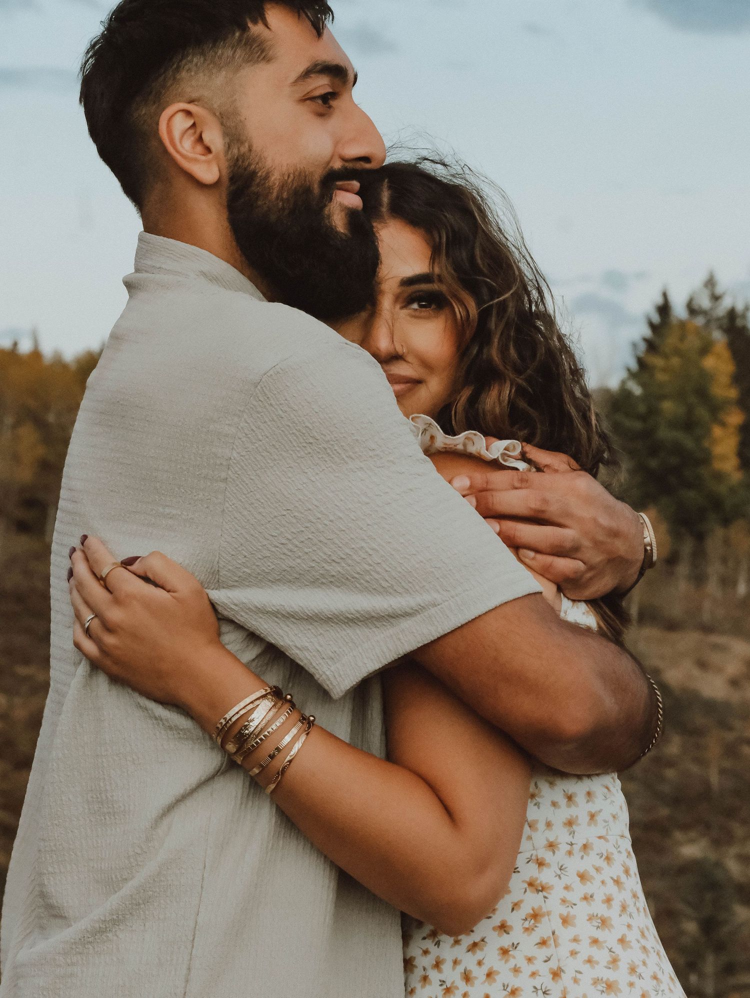 Couple embracing outdoors in autumn, woman in floral dress with gold bracelets hugging bearded man in beige shirt.