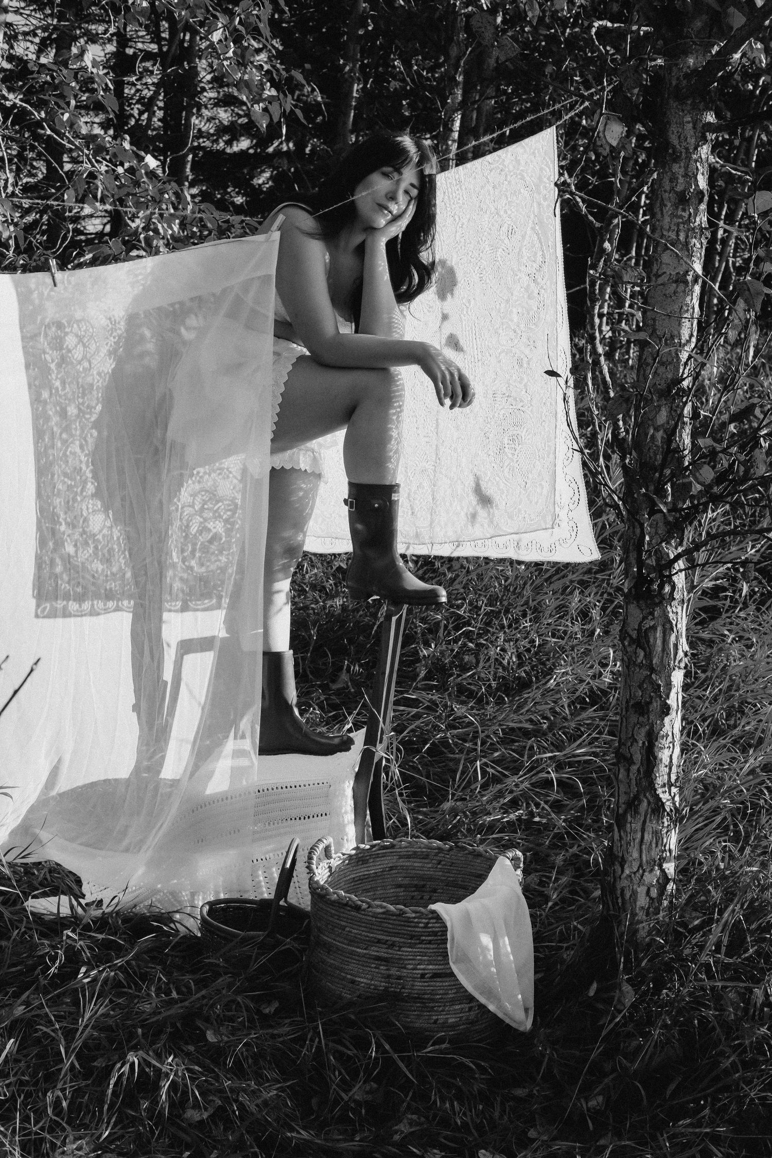 Black and white photo of a woman in boots posing by a clothesline with hanging laundry outdoors.