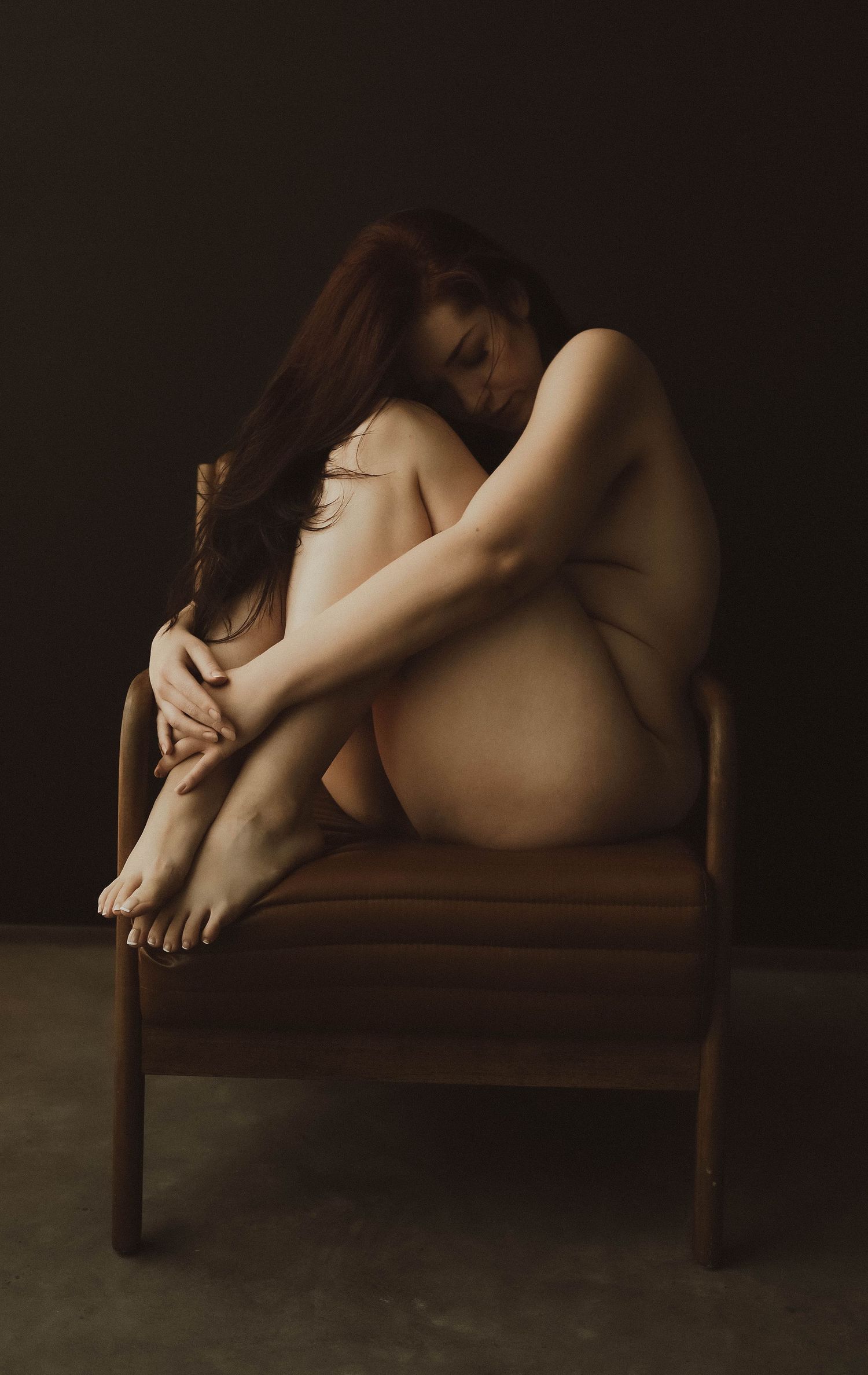 A nude woman sits curled on a wooden chair, hugging her knees in a moody, dark studio setting.