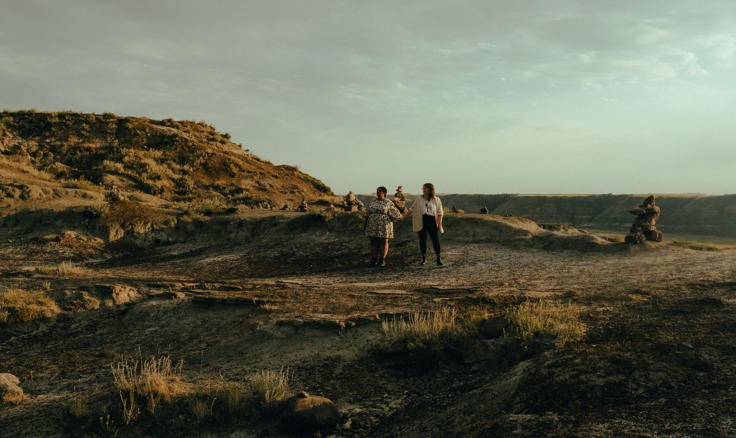 Two people walking hand in hand across a vast dark volcanic landscape under a cloudy sky.