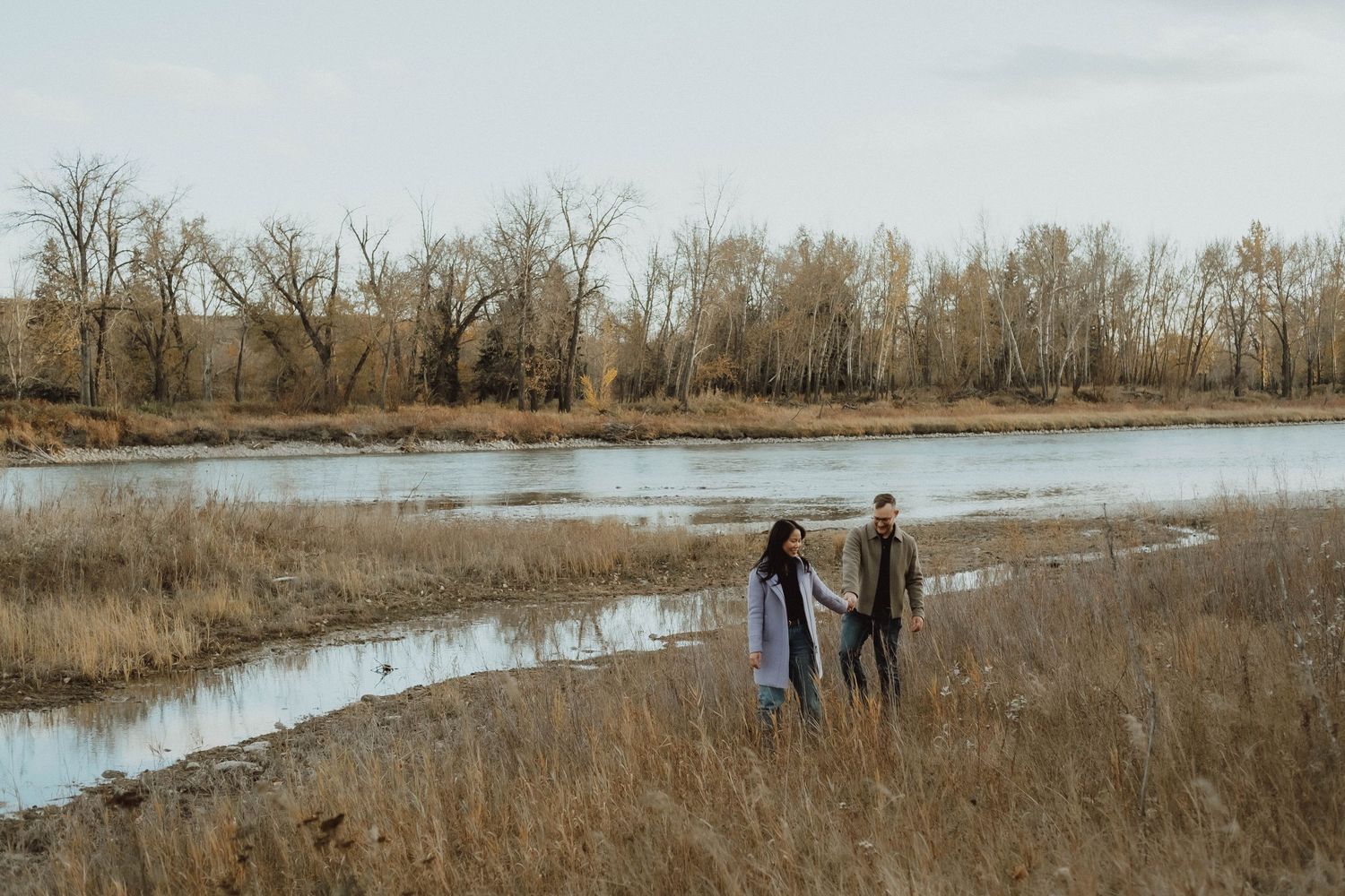 Couple holding hands walking along a serene river bank surrounded by golden grass and bare trees in autumn.
