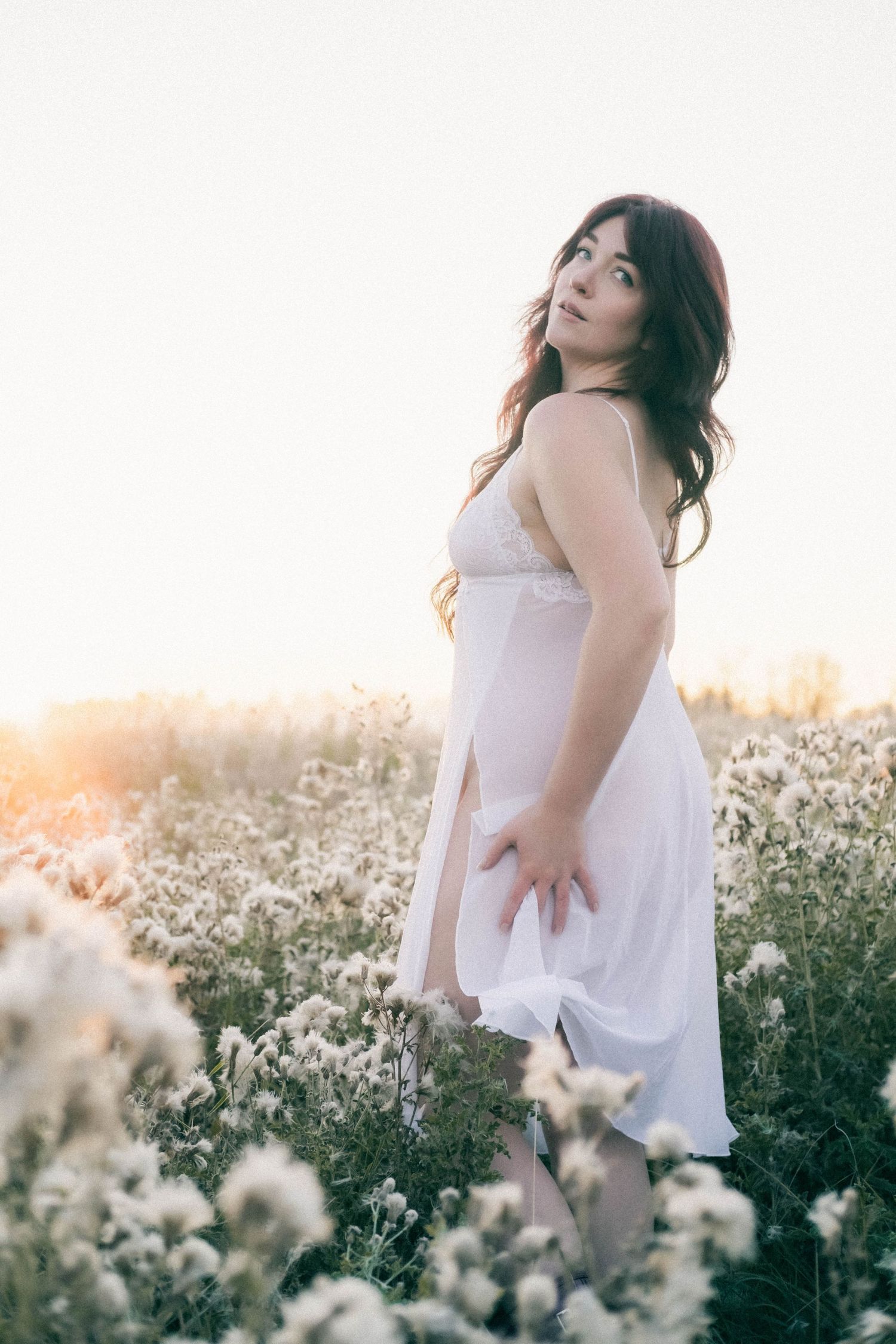 Woman in white dress standing in wildflower field at golden hour sunset.