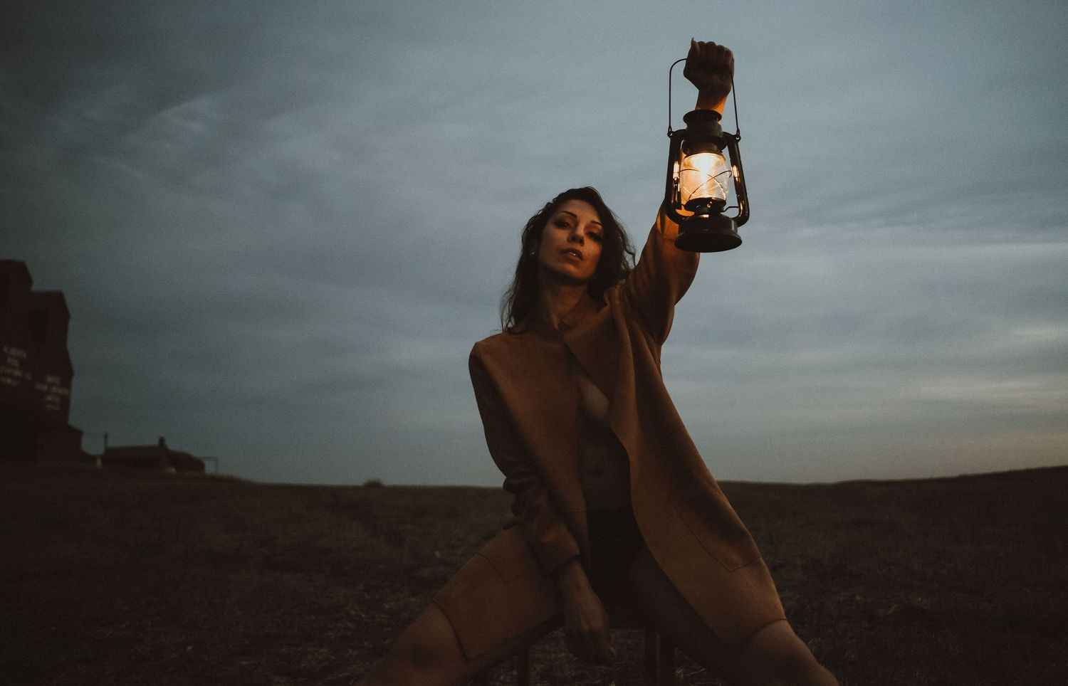 Woman in beige coat holds glowing vintage lantern aloft in dramatic desert landscape under moody twilight sky.