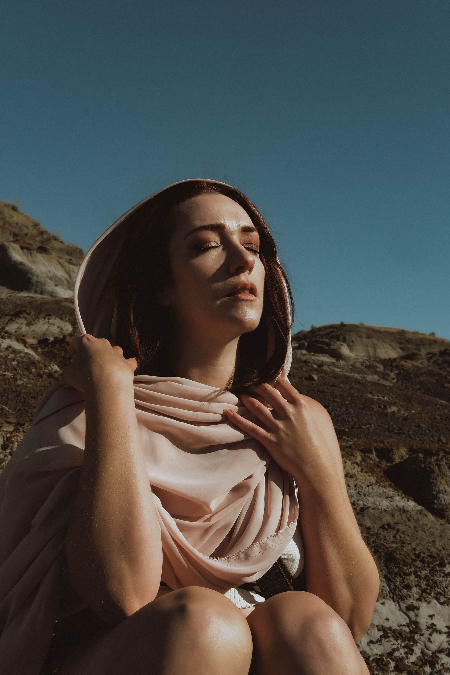 Woman in pink hood sits against rocky terrain, eyes closed, basking in sunlight under a clear blue sky.