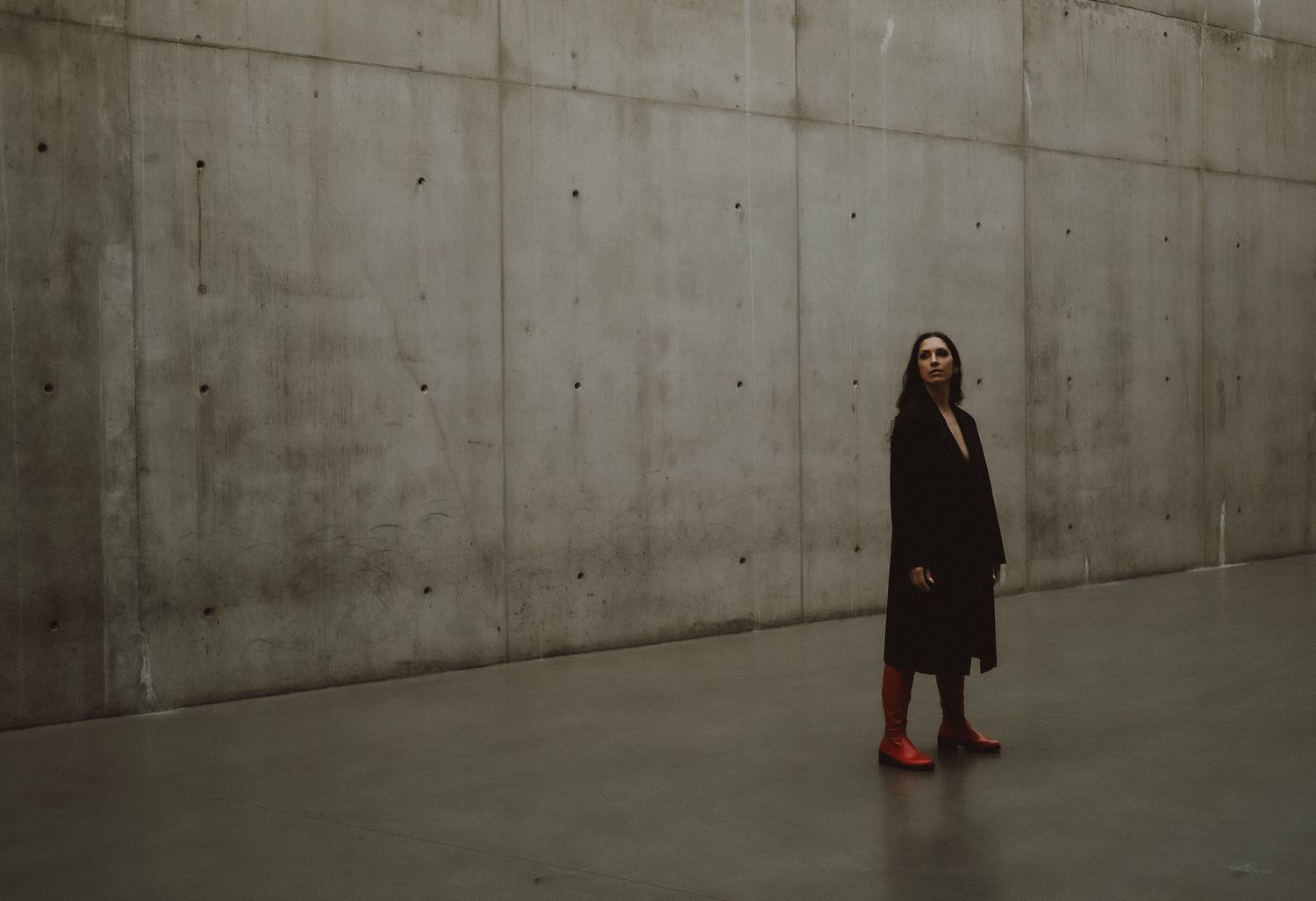 Woman in black coat and red boots standing in a minimalist concrete space, looking upward.