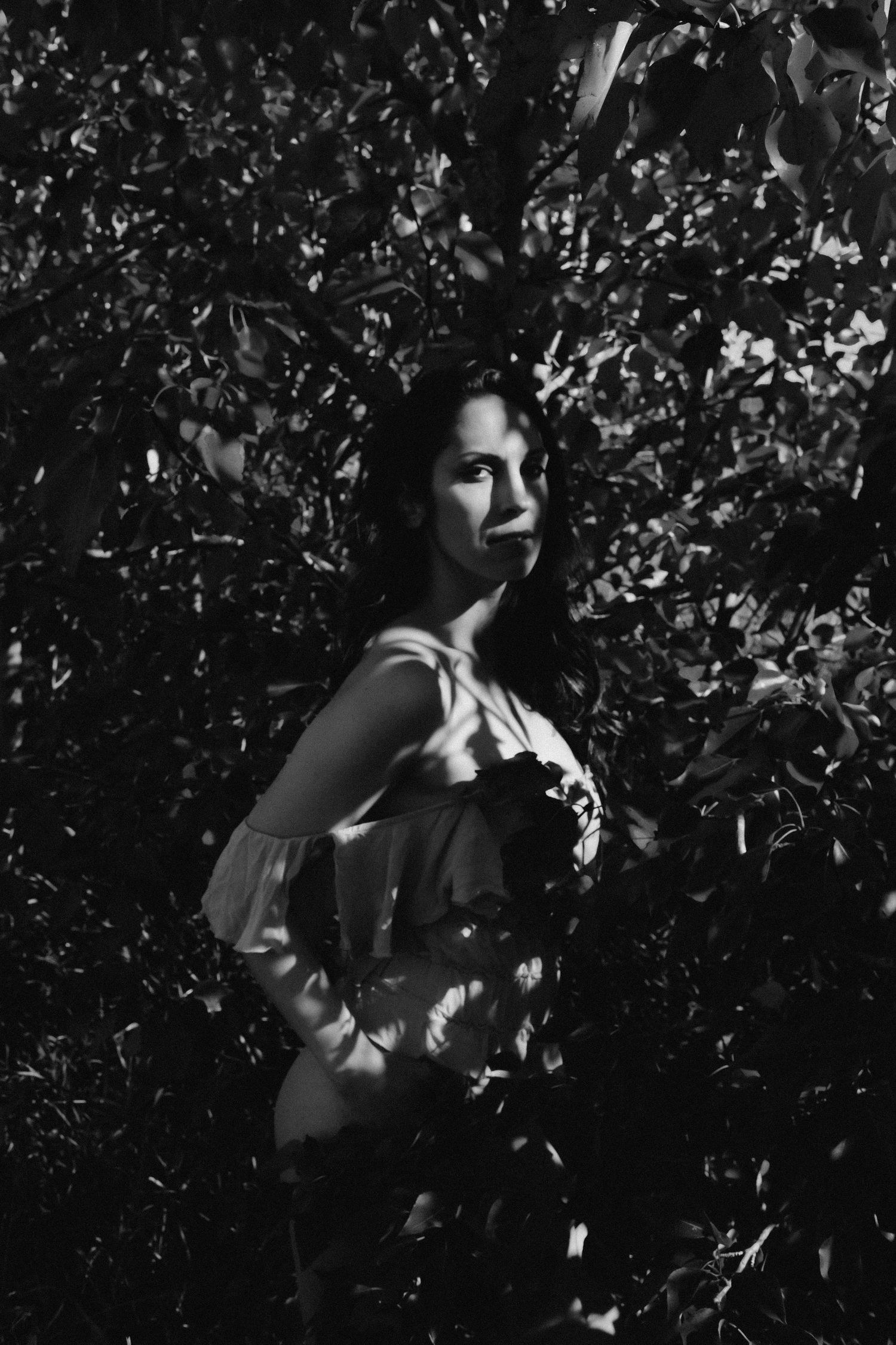Black and white portrait of a woman standing among lush leafy bushes, with dramatic light and shadow contrasts.