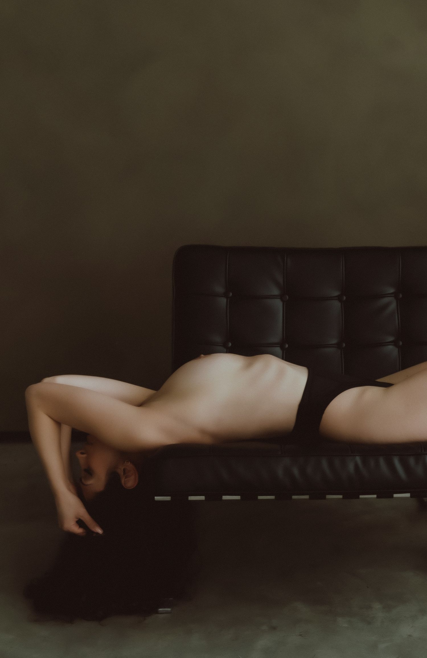A nude figure reclining gracefully on a dark leather chaise lounge against a moody olive-green background.