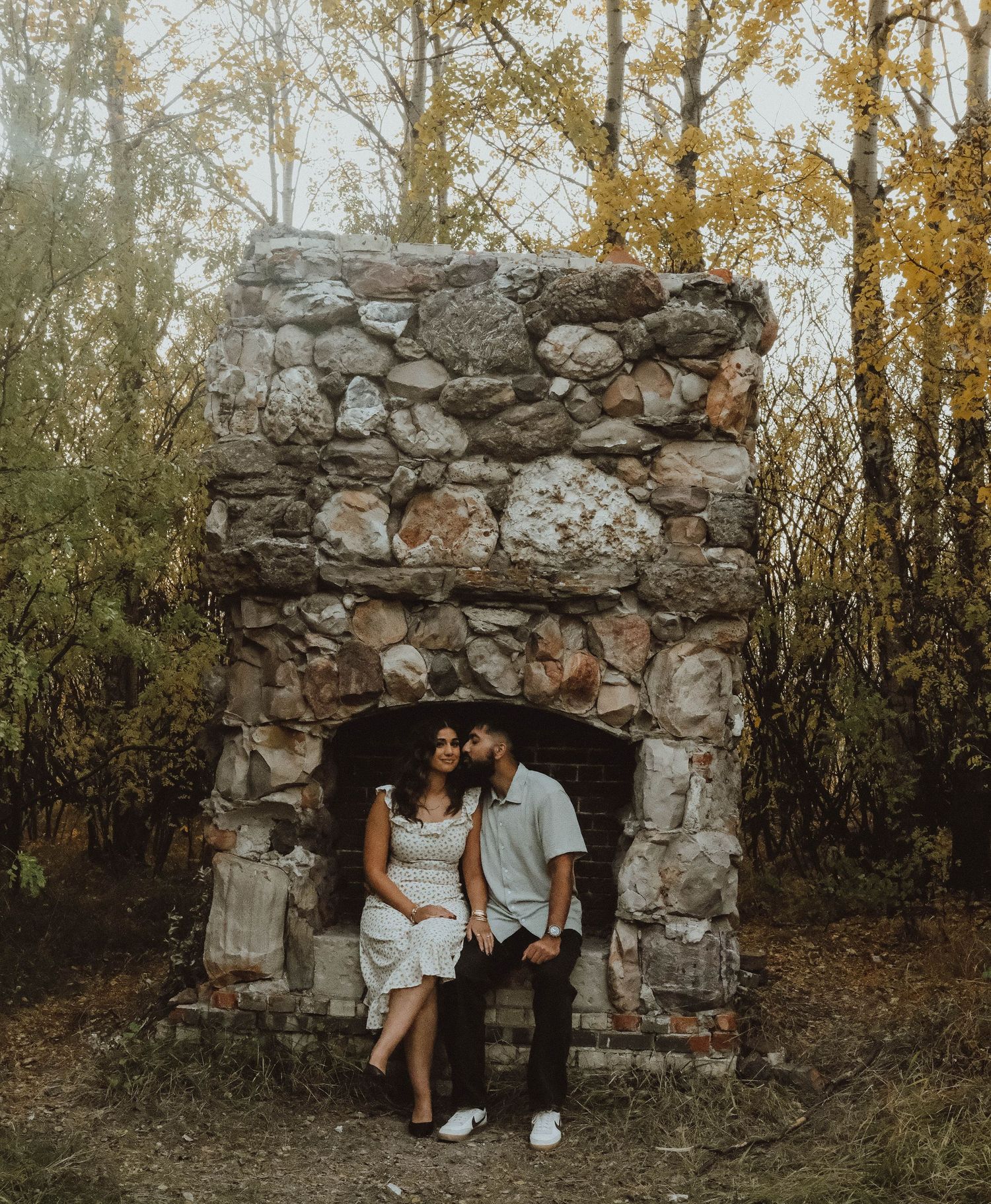 Couple sitting together in a rustic stone fireplace outdoors surrounded by autumn trees in a romantic engagement photo.