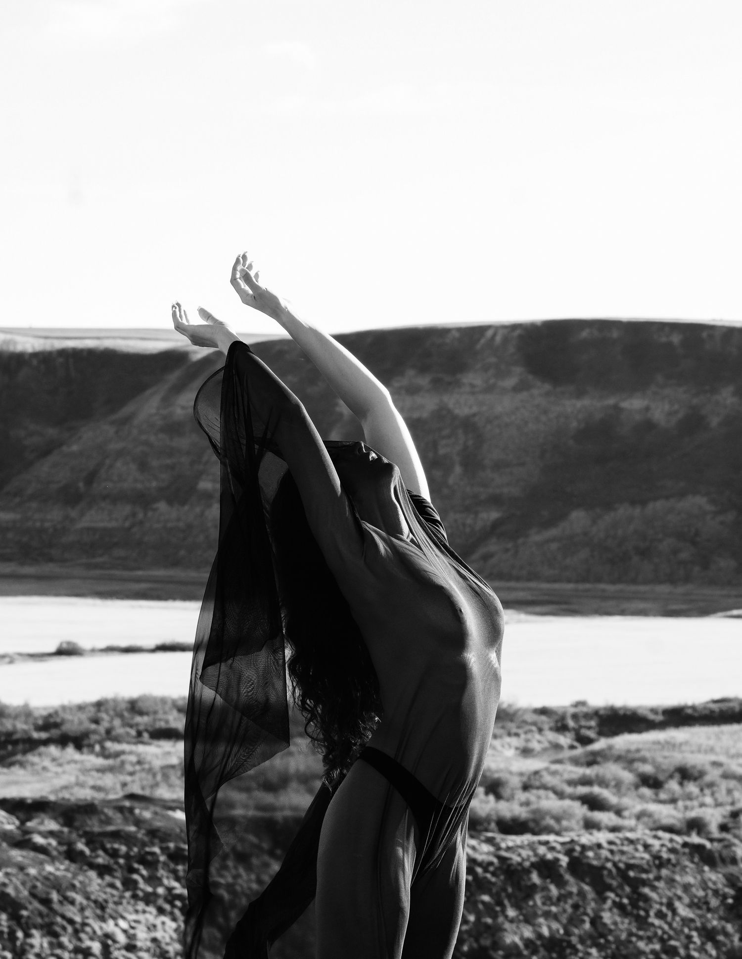Black and white photo of a dancer in sheer fabric, arms raised gracefully against a scenic landscape with hills and water.