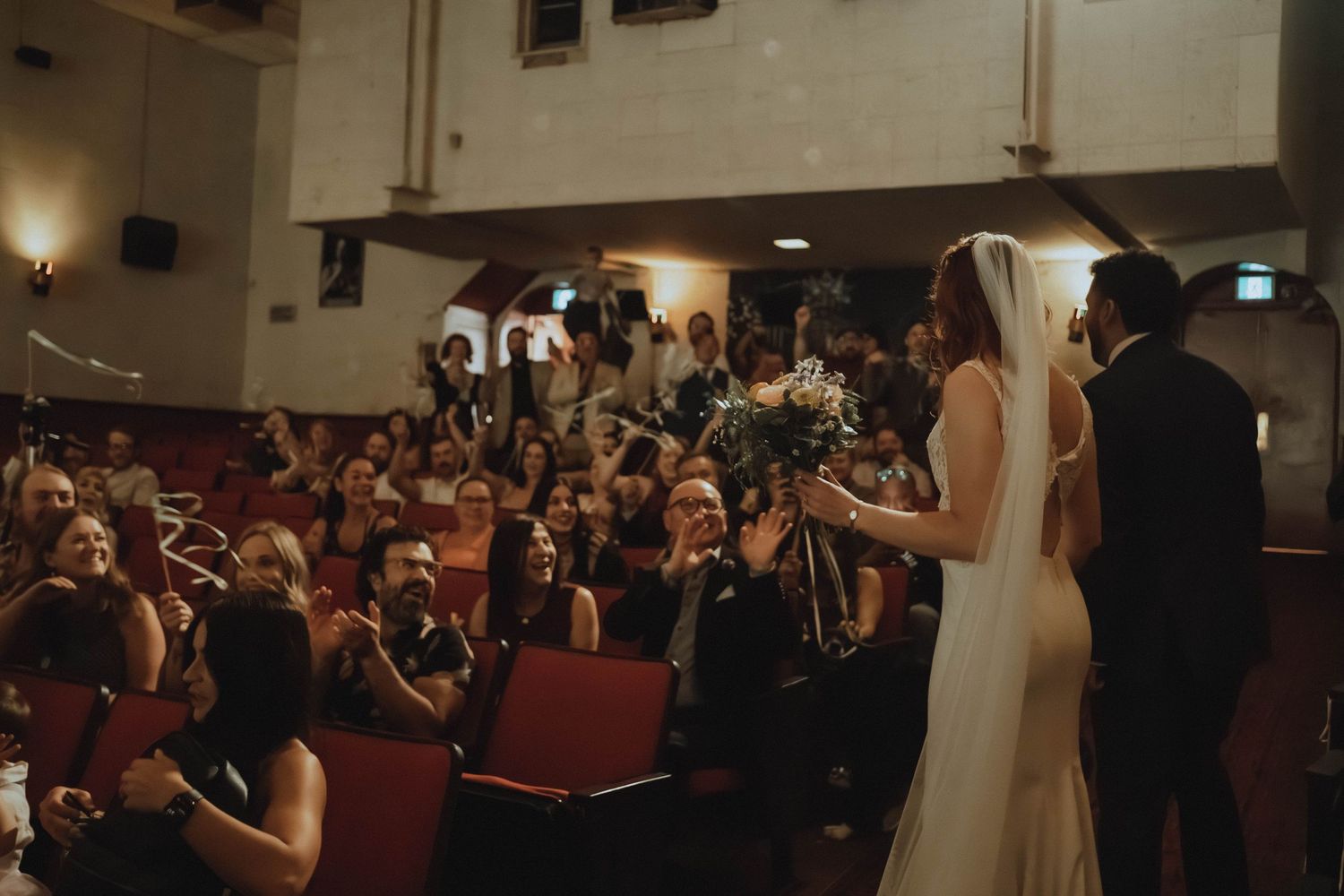 Bride and groom stand at the altar as wedding guests celebrate in a beautifully decorated theater venue.
