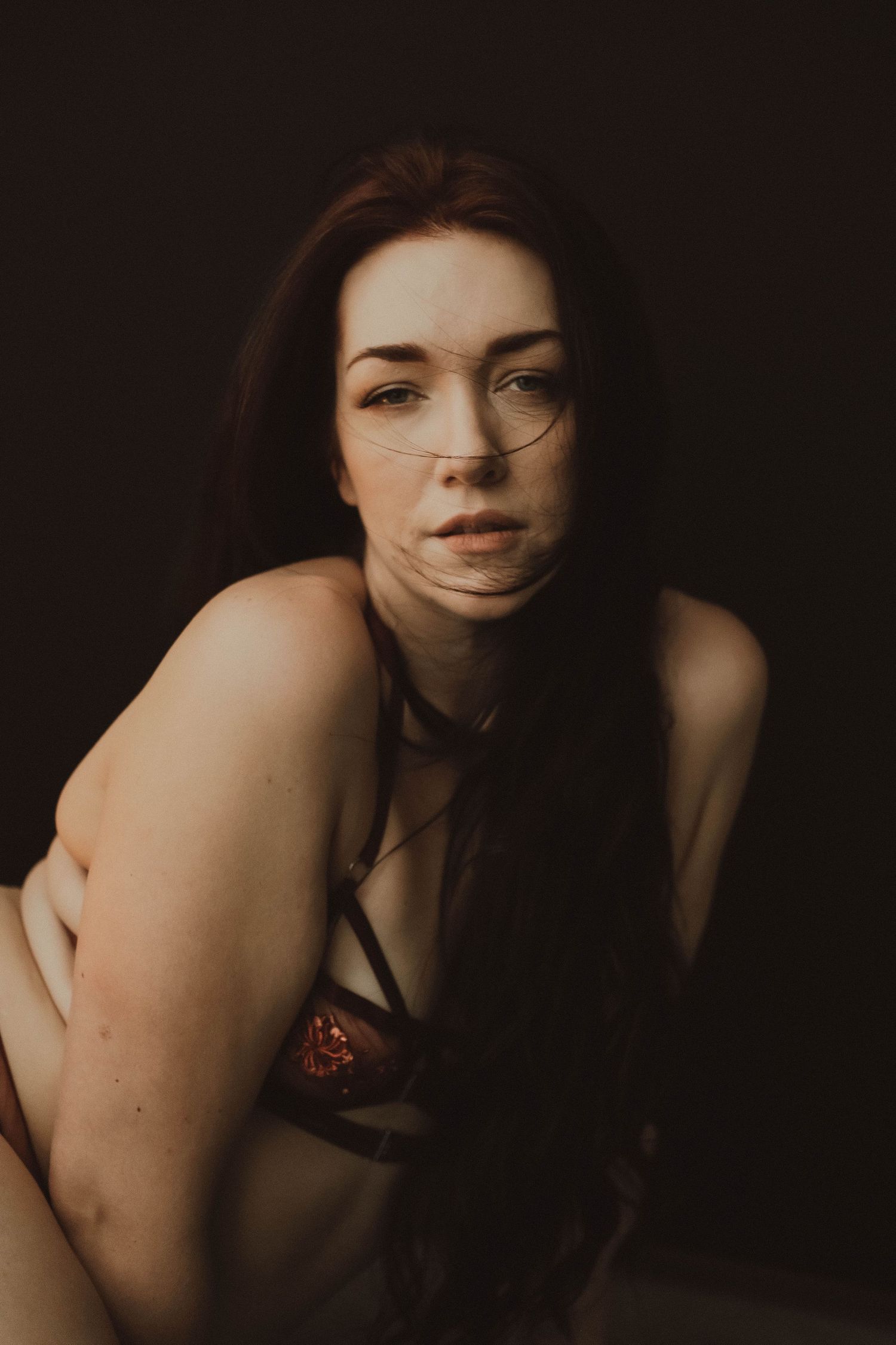 A woman with red hair and glasses poses in moody dark studio lighting wearing burgundy lingerie.