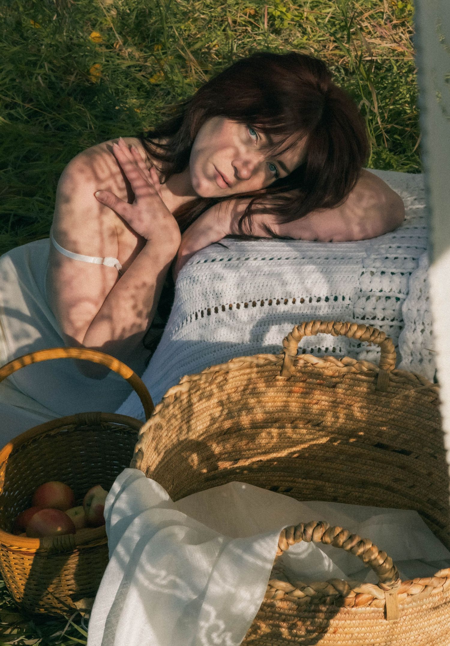 Woman resting on white blanket outdoors beside wicker baskets with apples, dappled sunlight on skin.