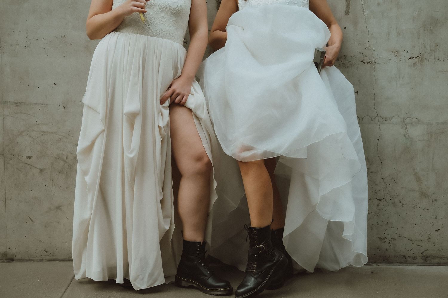 Close-up of two women in white wedding gowns and brown boots standing against a concrete wall.