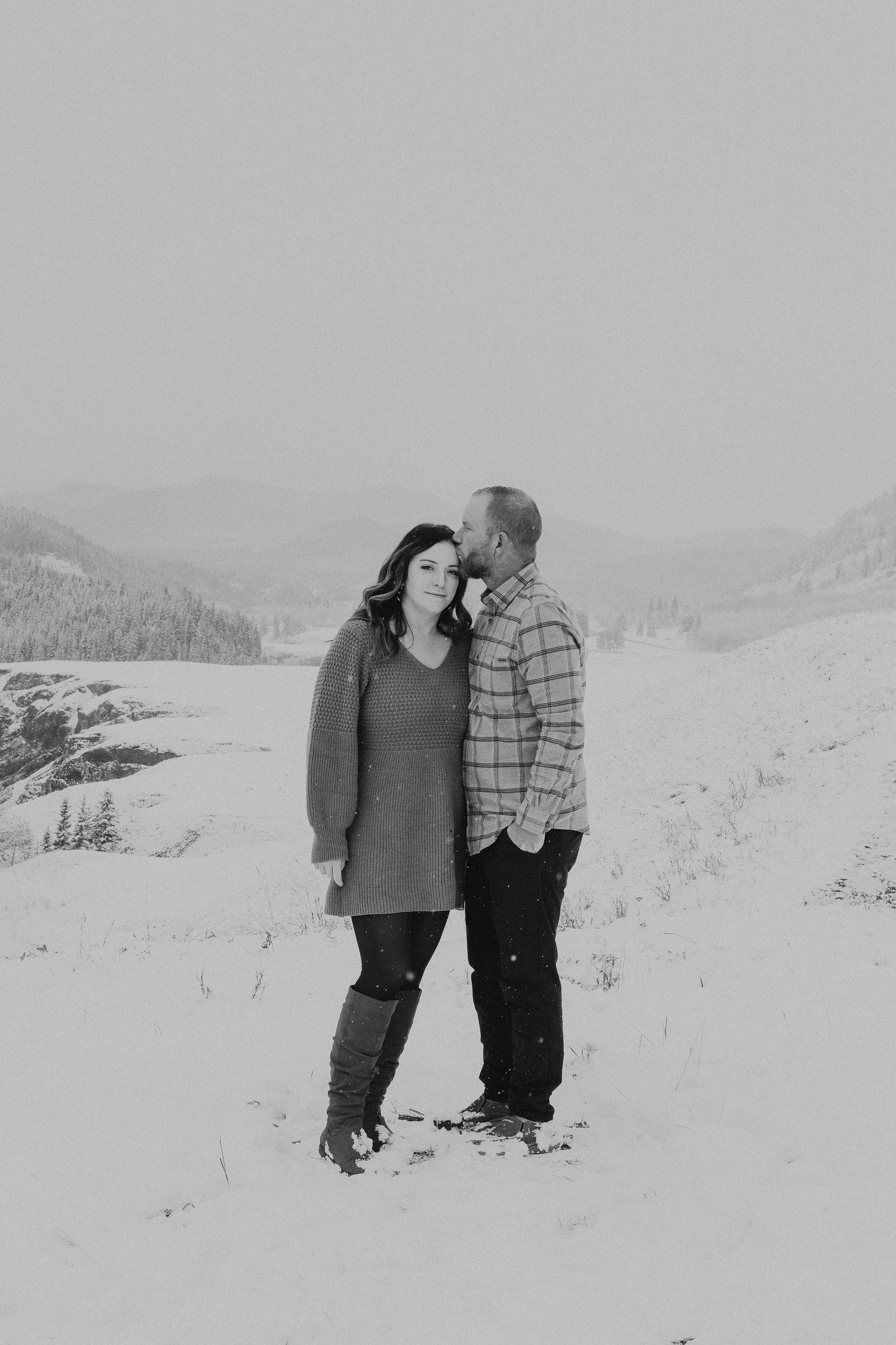 Black and white photo of a man kissing woman on cheek in snowy landscape.