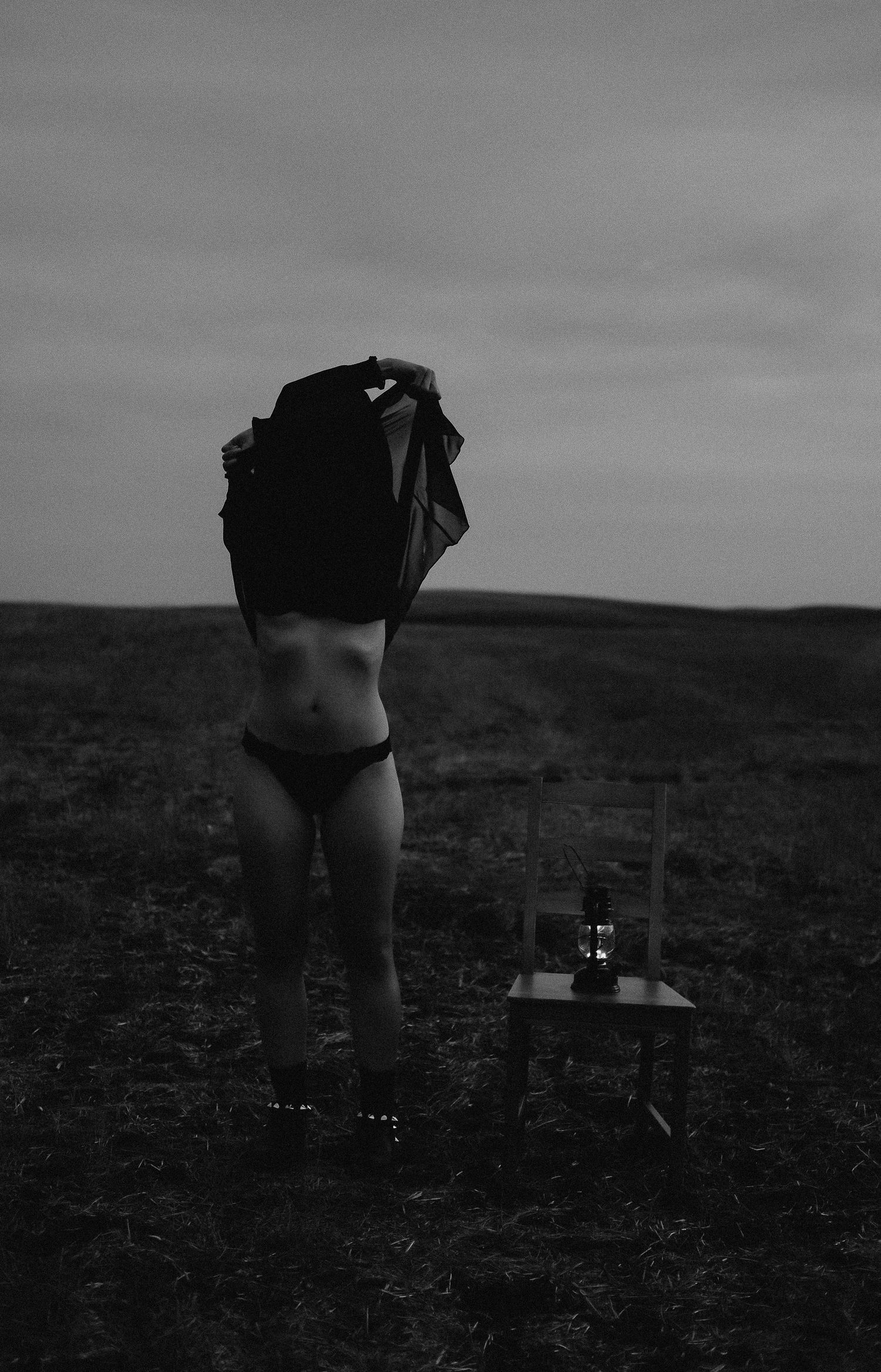 Black and white photo of a woman removing her jacket in an open field at dusk, with a small table beside her.