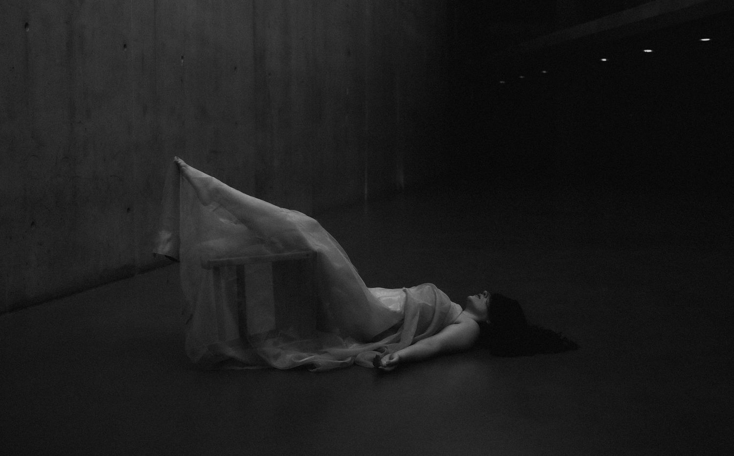 Black and white photo of a person lying on a dark floor draped in white fabric in a minimalist space.