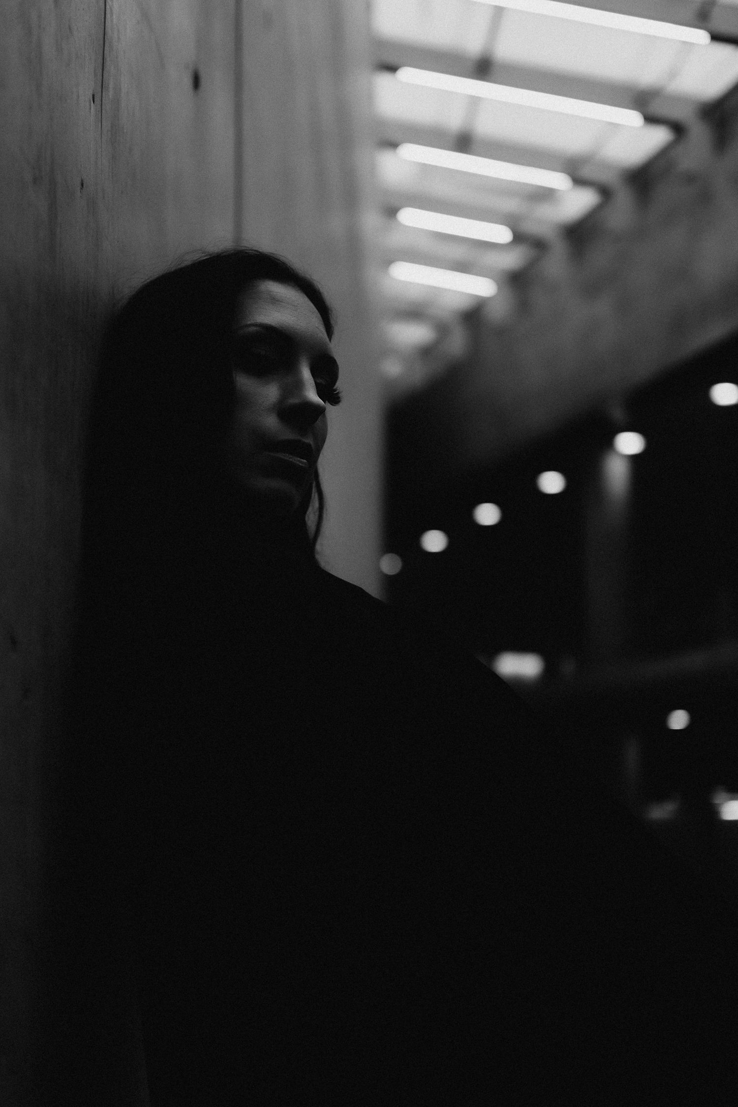 Black and white portrait of a woman leaning against a wall in a dimly lit corridor with bokeh lights.