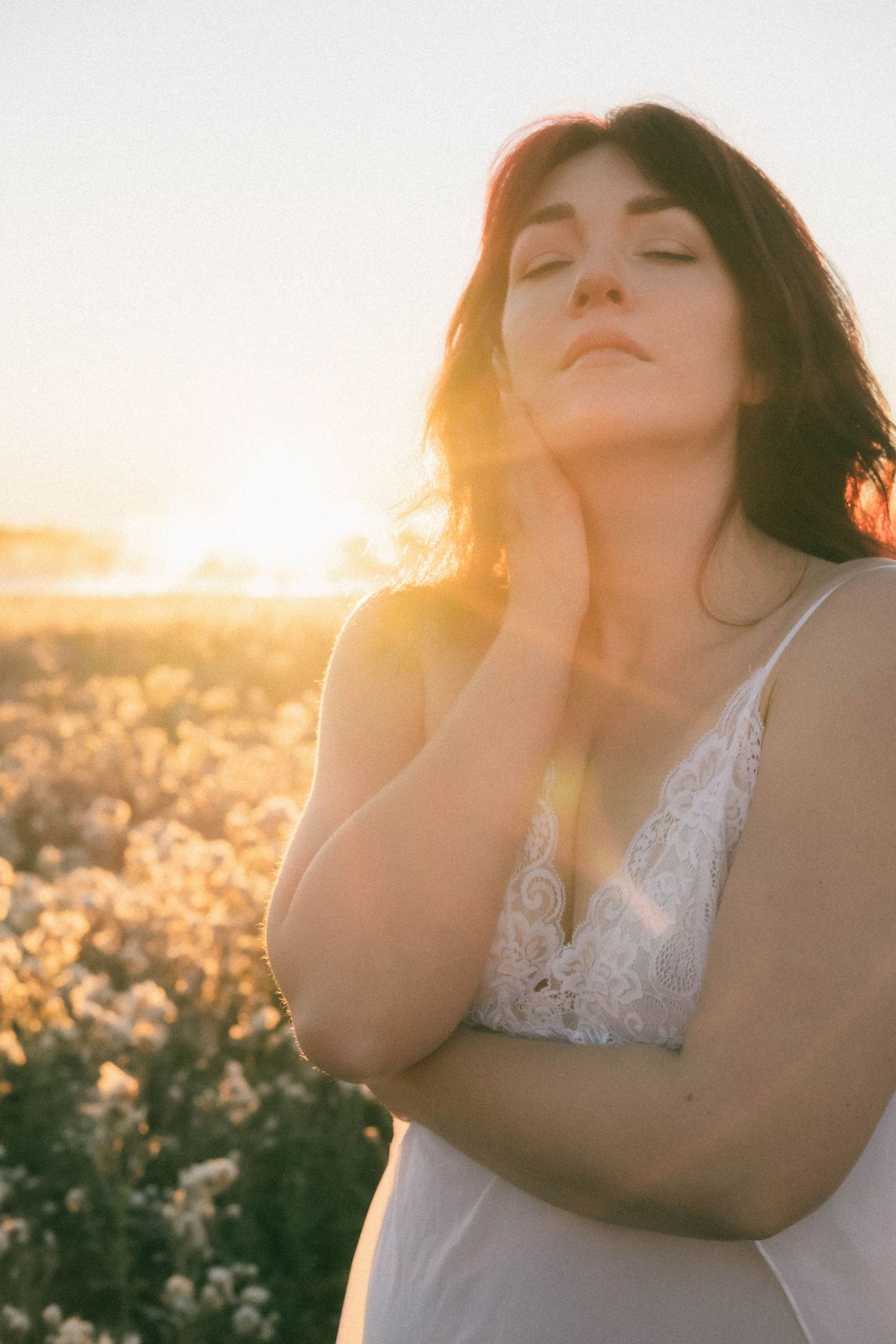 Woman in white lace dress standing in flower field at golden sunset with eyes closed and hand raised to face.