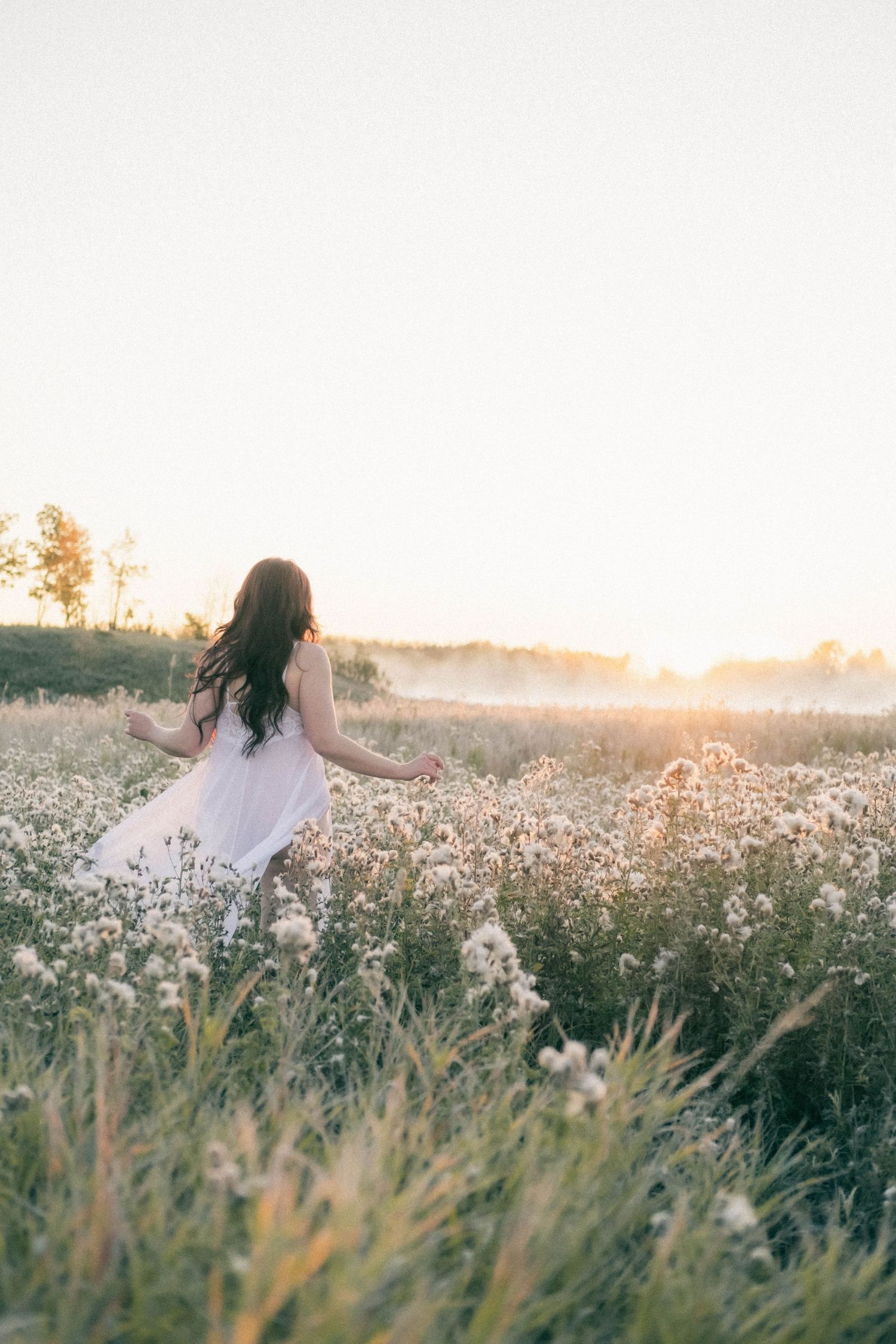 A woman in a white dress walking through a misty wildflower field at golden hour sunset.