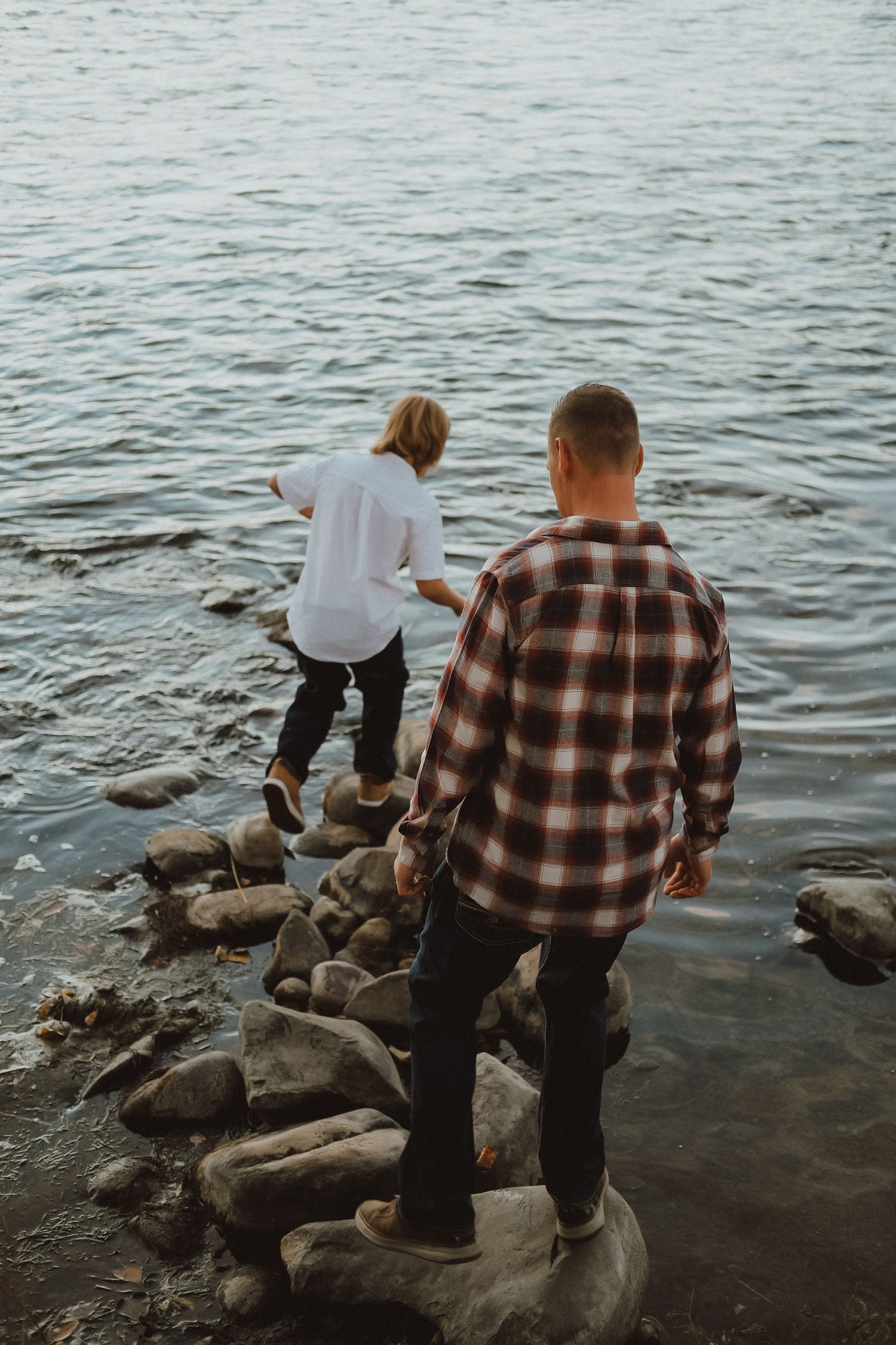 Father and son stepping across rocks along a serene lakeside shore during golden hour.