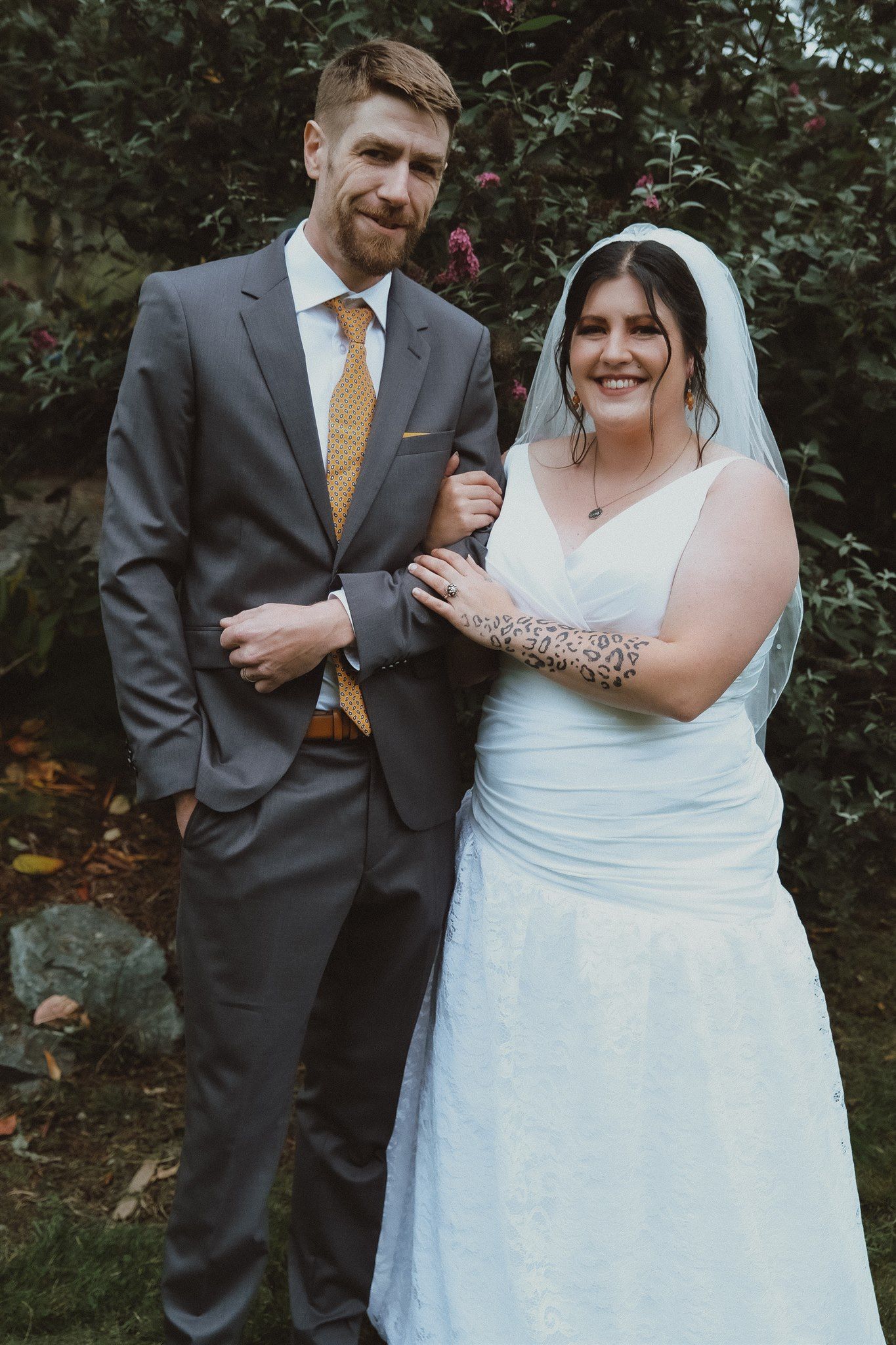 Bride and groom posing outdoors, she wearing a white gown with veil and leopard print tattoo, he in a gray suit.