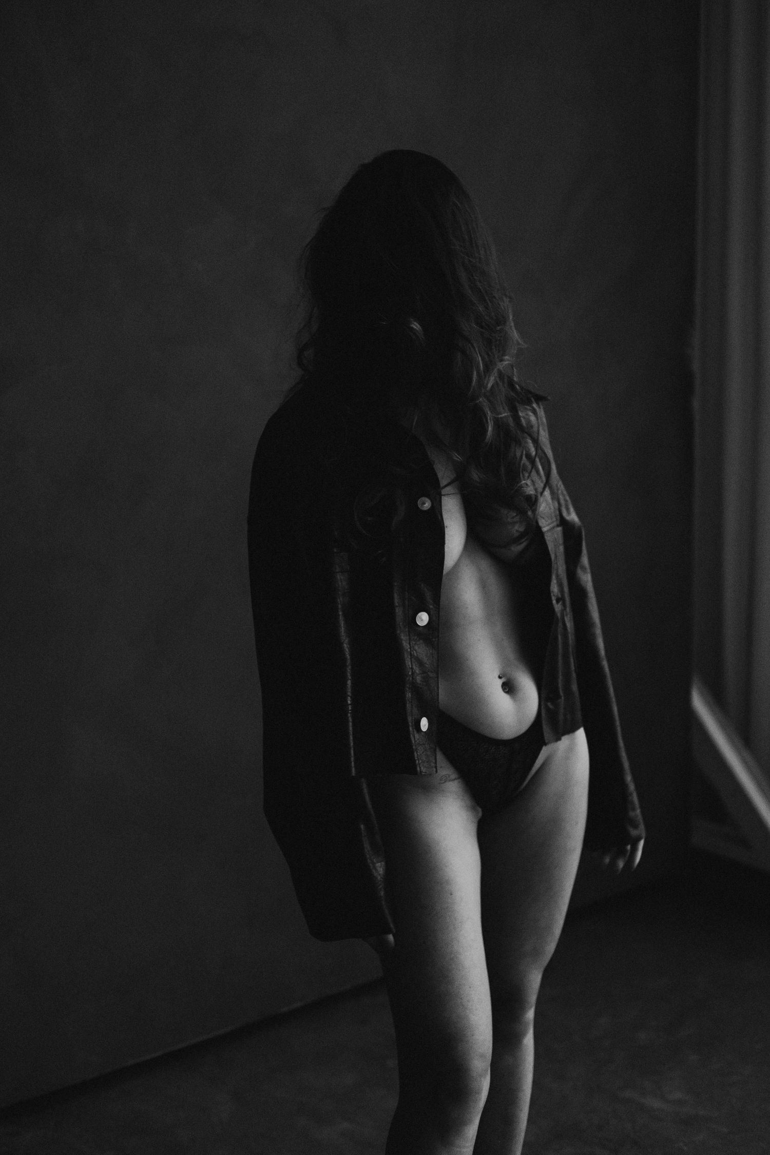 Black and white photo of a woman in an open denim jacket and underwear standing in a dark studio.