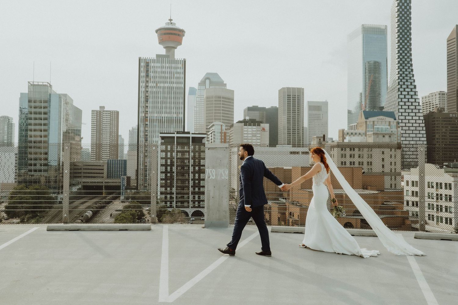 Bride and groom hold hands walking on rooftop with Vancouver city skyline backdrop on their wedding day.