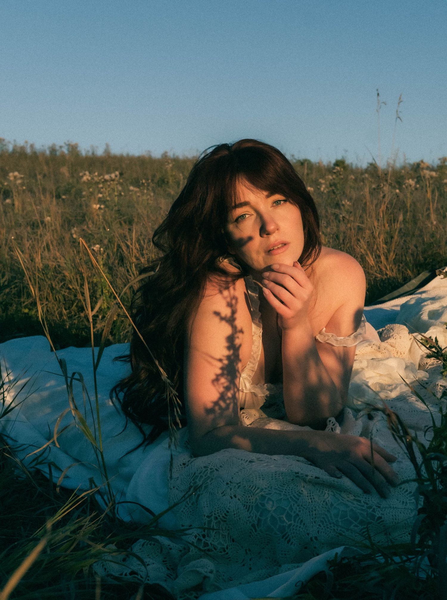 Young woman with long dark hair lying in a wildflower field, wearing a white lace dress in golden hour light.