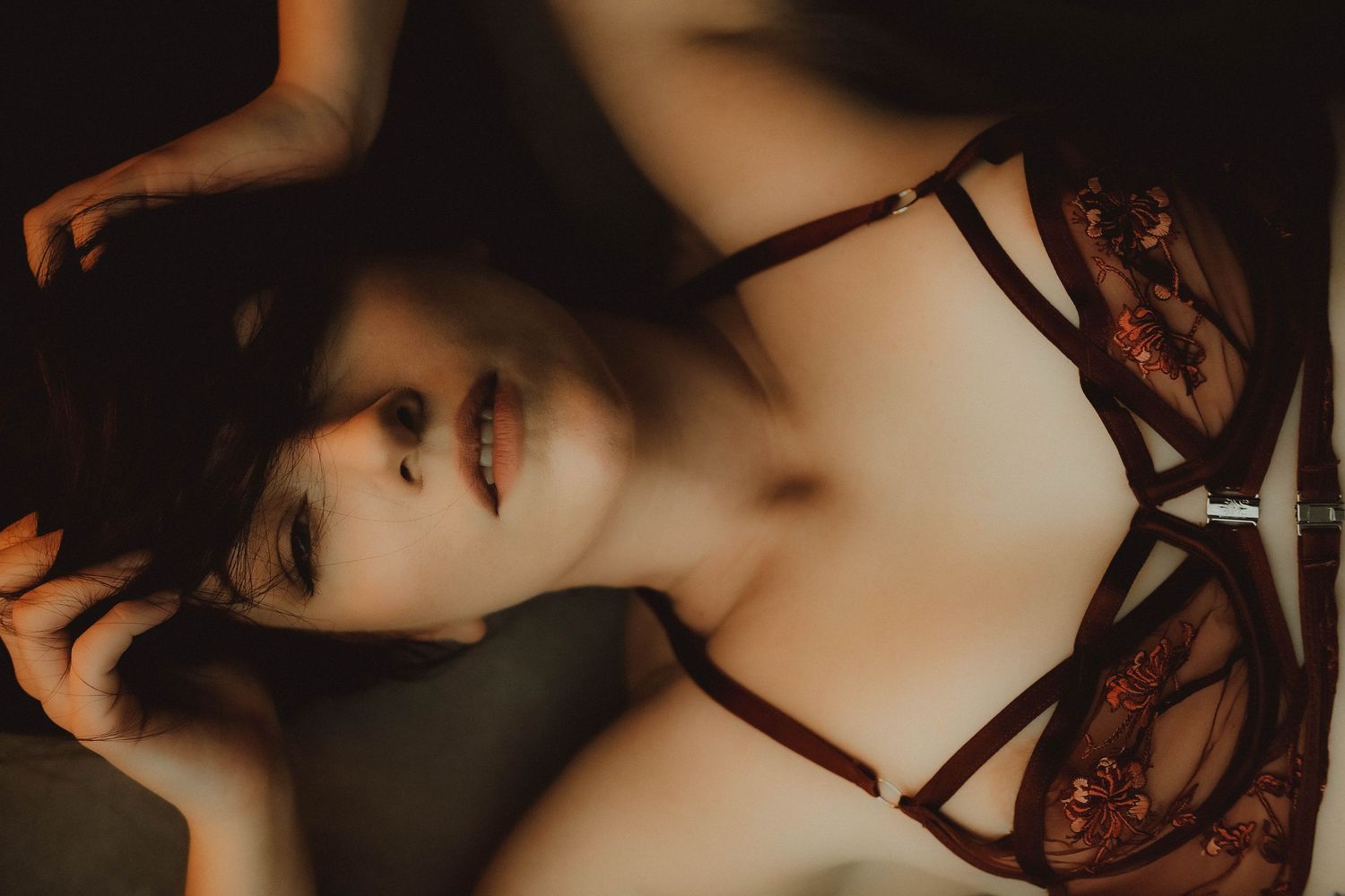 A woman in elegant dark red lace lingerie reclines gracefully in soft, moody lighting.