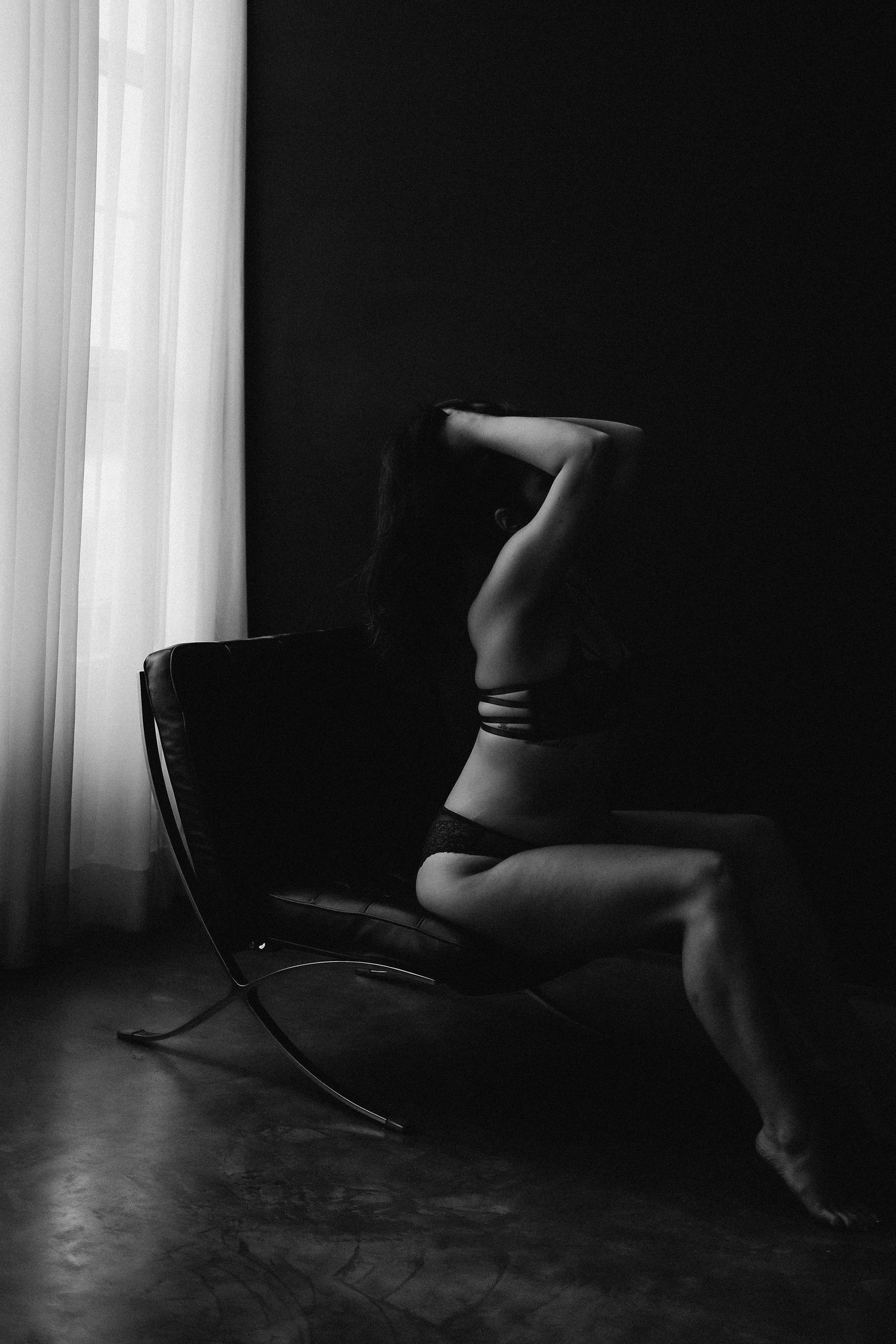 Black and white boudoir photo of a woman in lingerie posing gracefully on a modern chair near a curtained window.