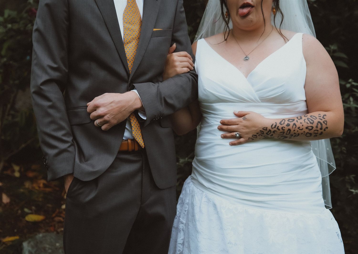 Bride and groom walking arm in arm outdoors, bride in white dress with leopard tattoo, groom in gray suit.