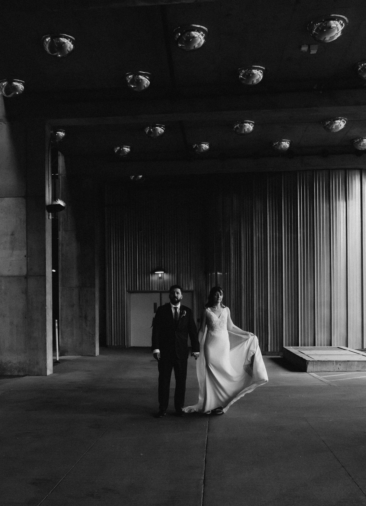 Black and white photo of a bride and groom holding hands in a dramatic architectural hallway with ceiling lights.