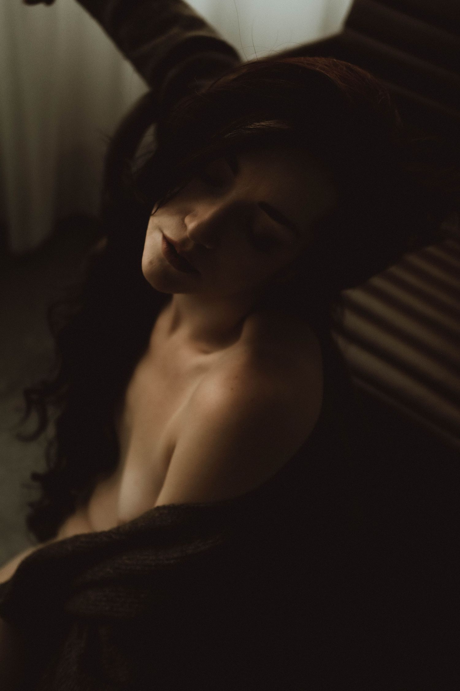 A woman with dark hair reclines in moody, dramatic lighting wearing an open robe in a sensual boudoir portrait.