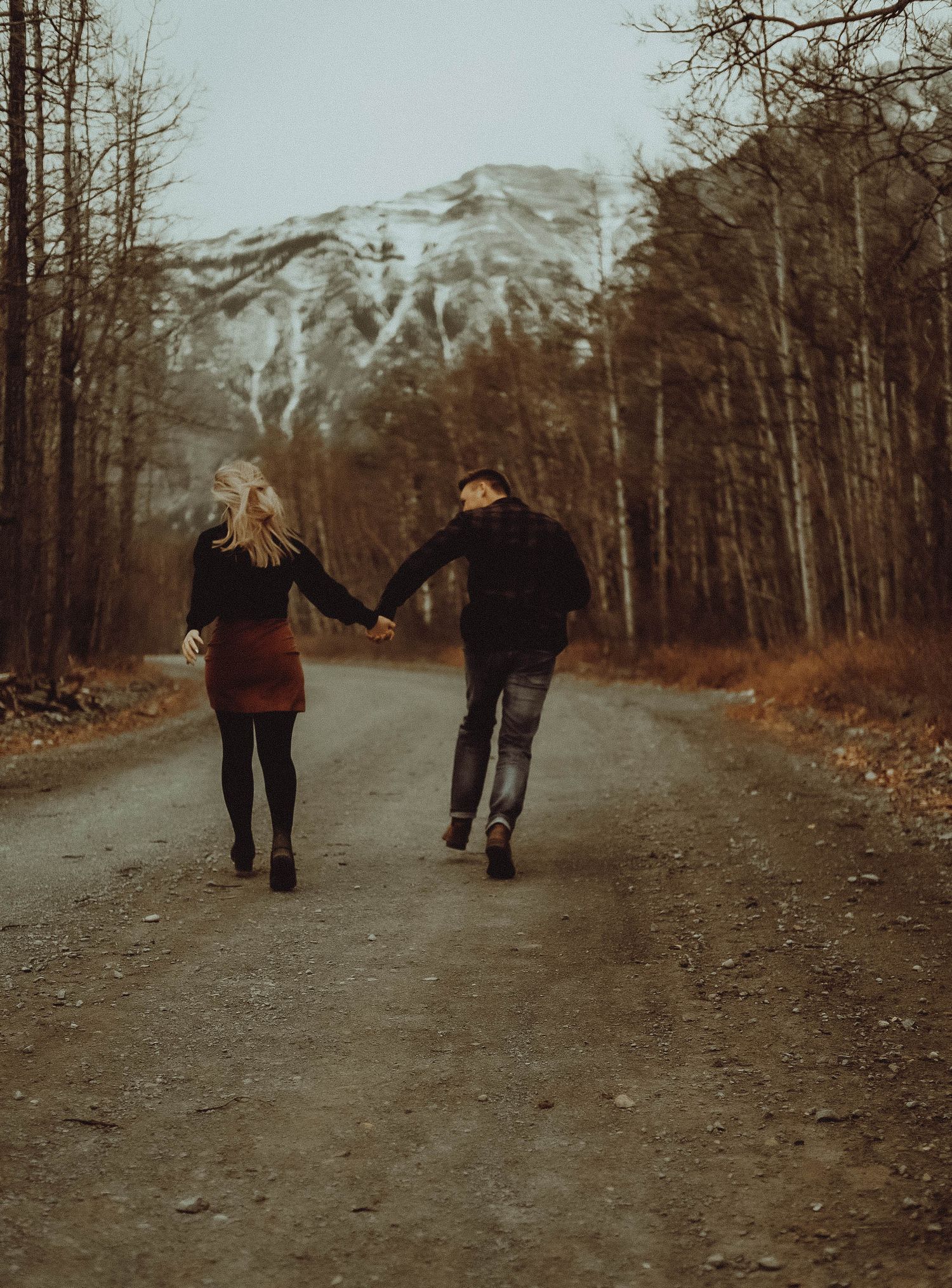 Couple holding hands walking down a scenic mountain road surrounded by bare trees and snow-capped peaks in autumn.