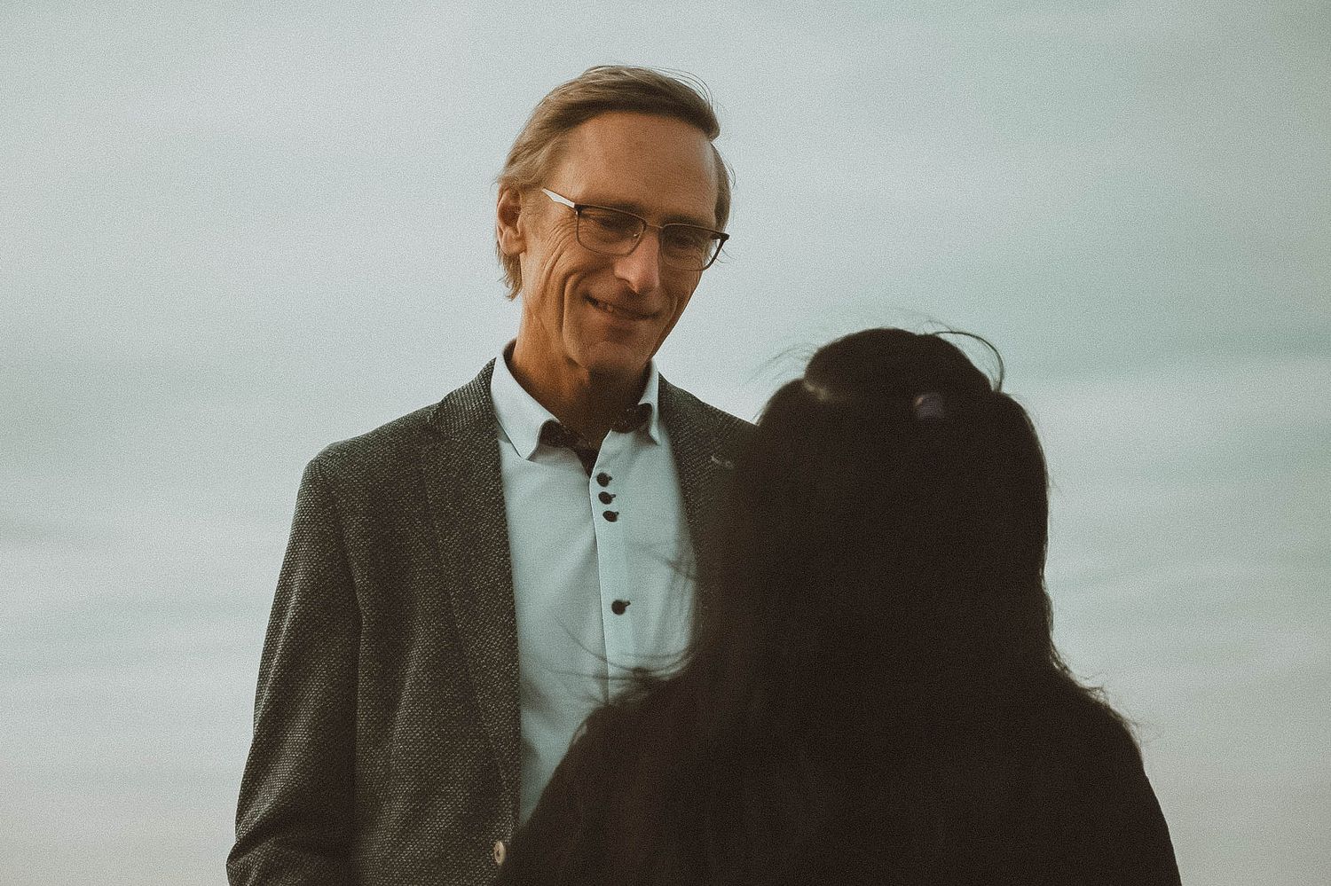 Smiling older man in glasses and gray blazer conversing with a dark-haired woman outdoors under cloudy skies.