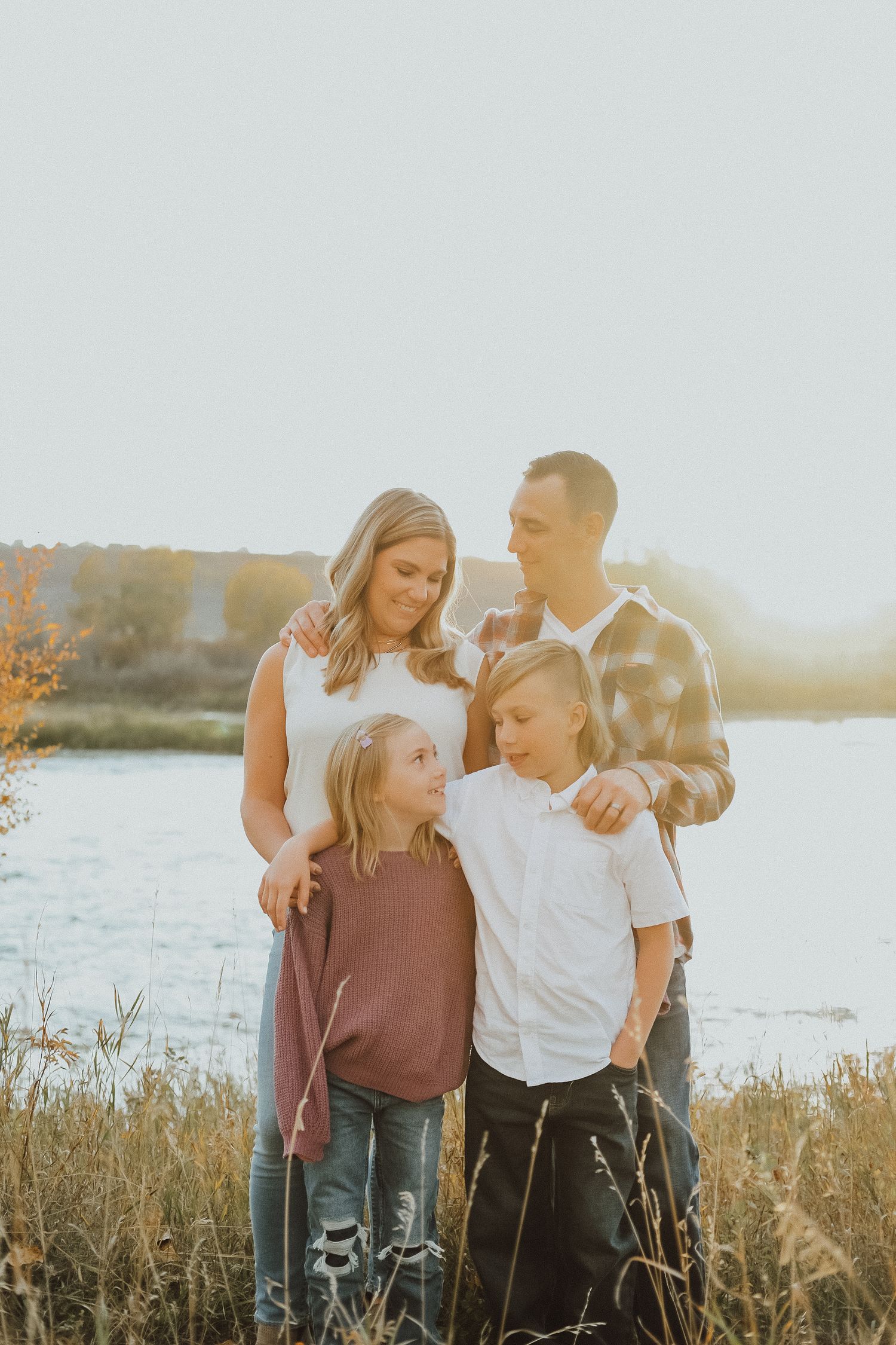 Family of four posing together in golden hour light near a lake surrounded by tall grass in autumn.