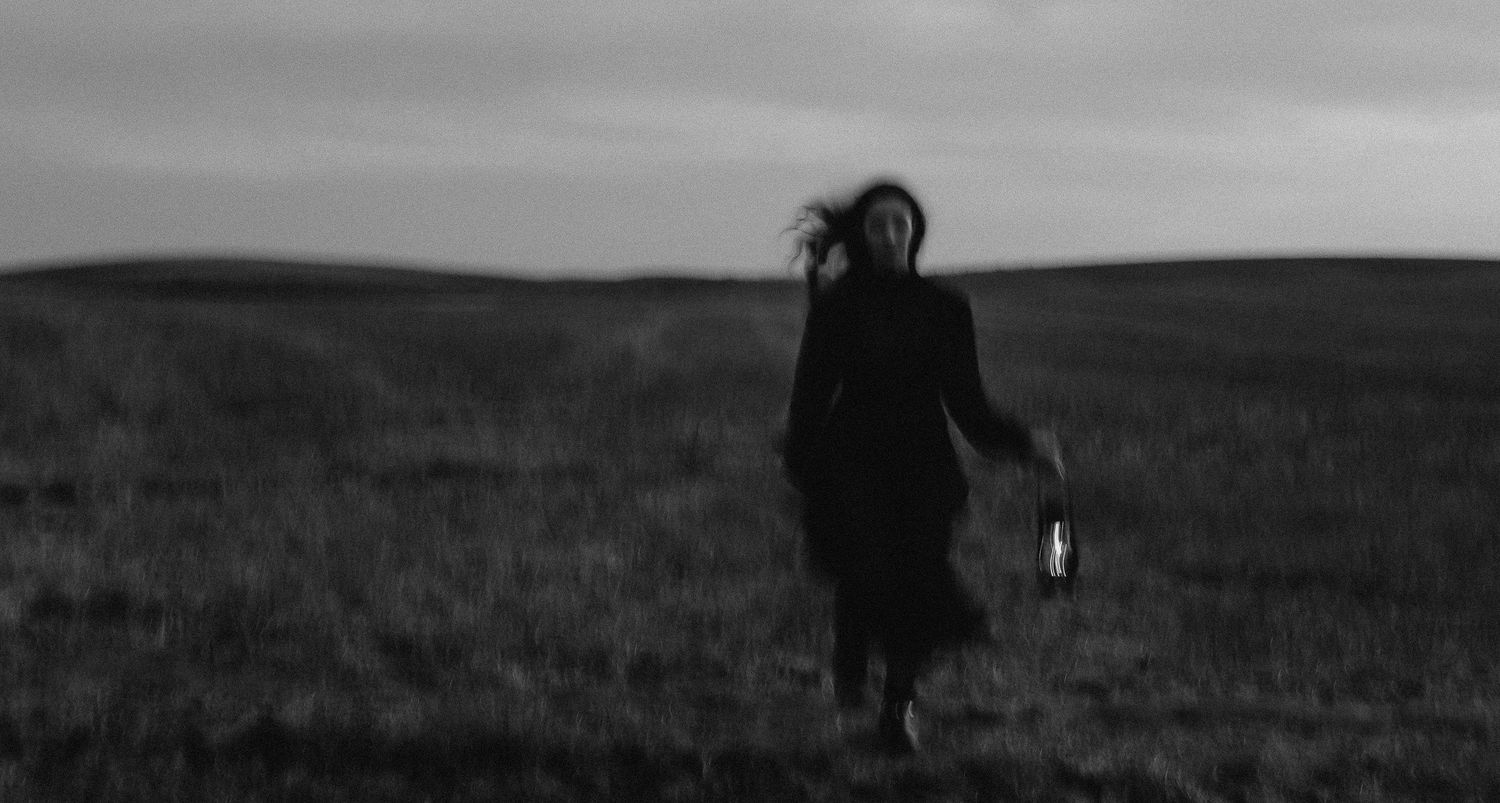 Black and white photo of a woman running through a field at dusk, holding a lantern, hair blowing in the wind.