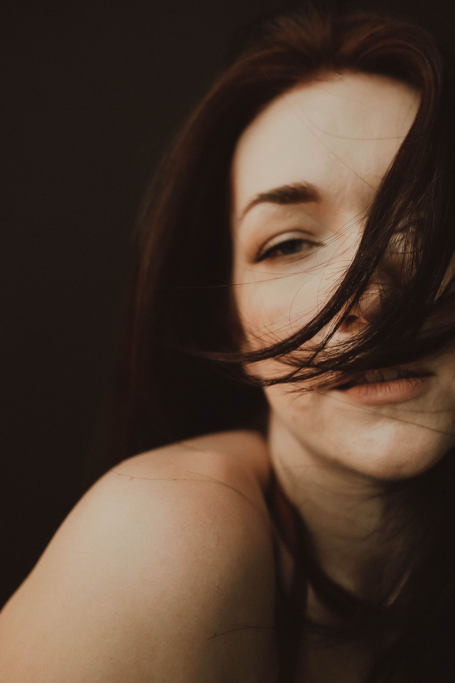 A young woman with windswept brown hair partially covering her face, smiling softly against a dark background.