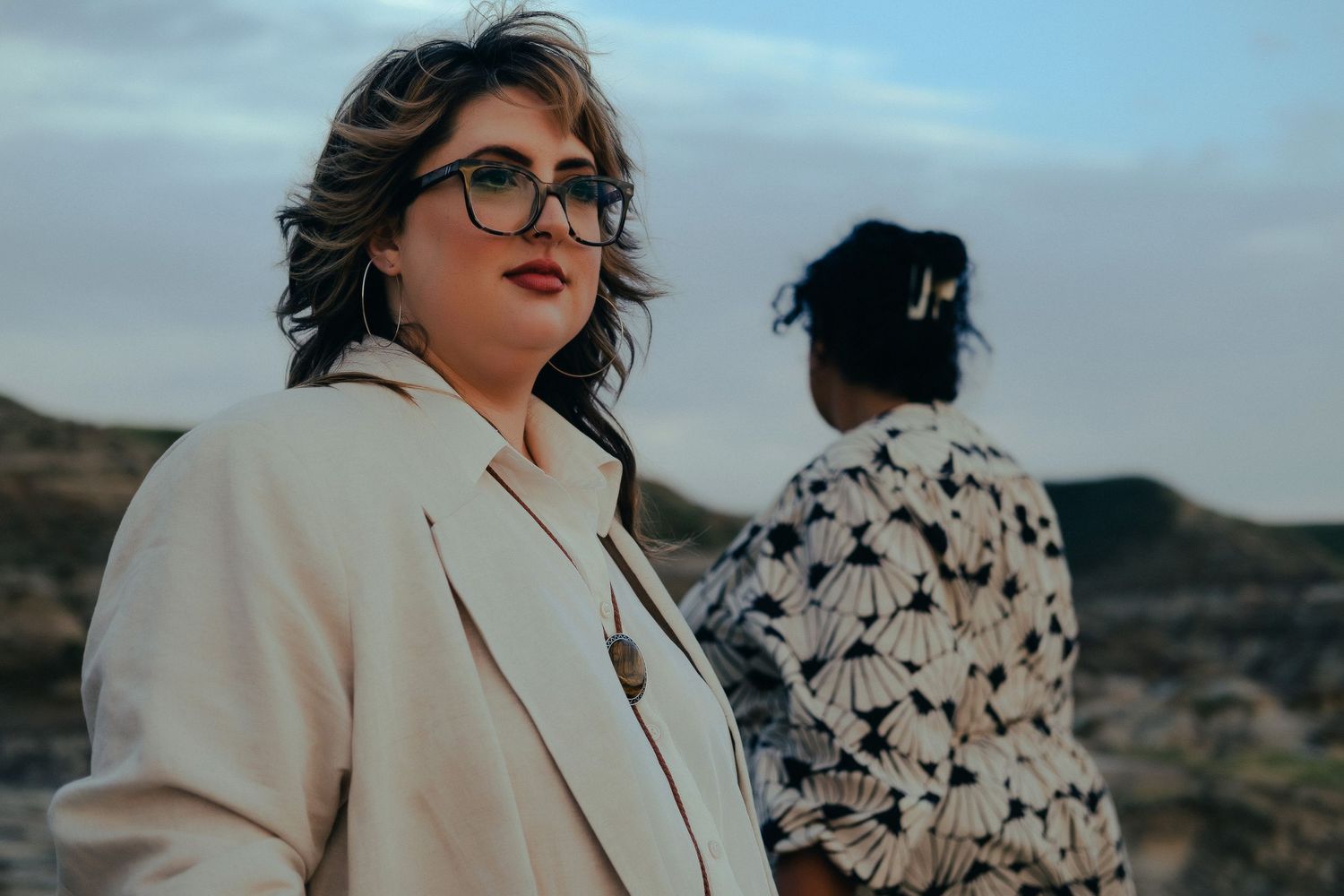 Woman in cream blazer with glasses stands confidently outdoors while partner stands softly behind her.