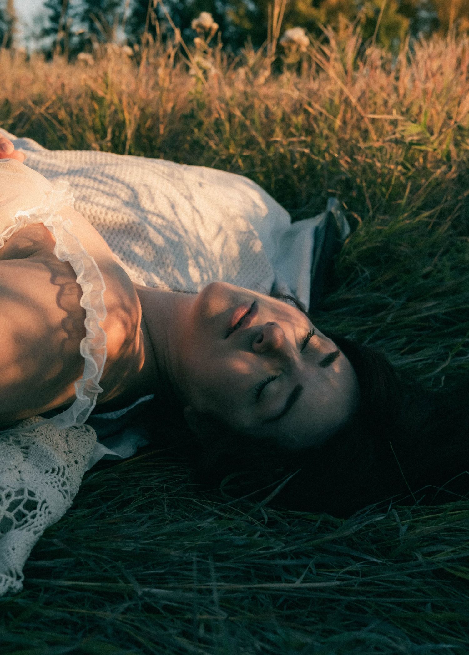 A young woman in a white lace dress lies peacefully in golden grass during sunset.