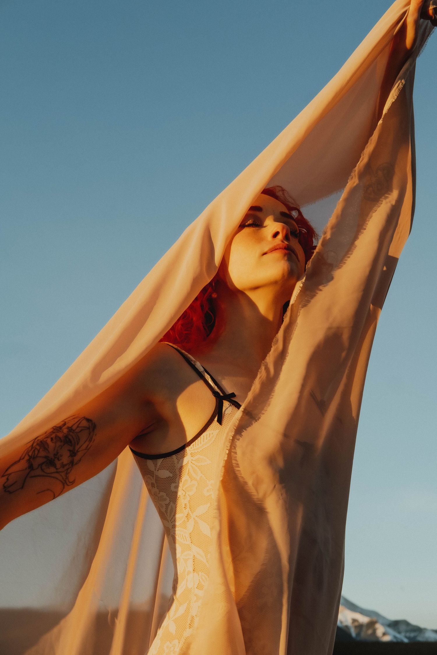 Woman with red headband and floral tattoo holds sheer fabric overhead against blue sky at golden hour.
