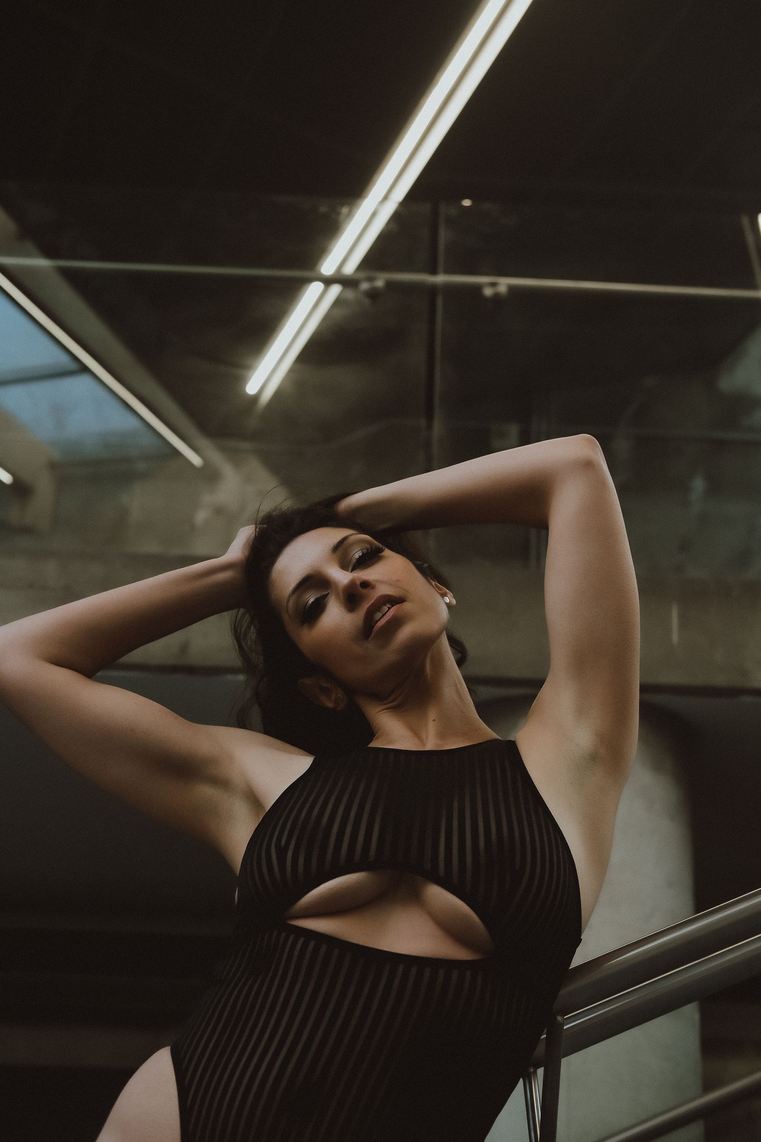 Woman in black ribbed cutout bodysuit poses with hands behind head near glass railing.
