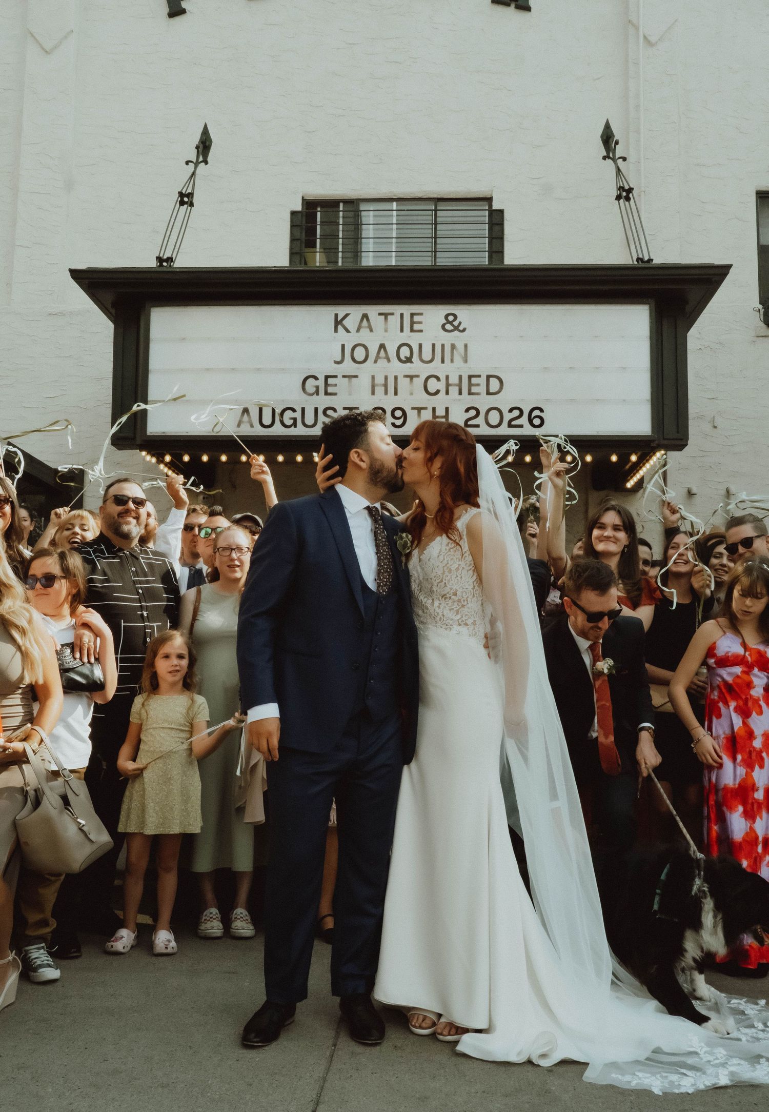 Bride and groom kiss outside venue under marquee sign reading Katie and Joaquin Get Hitched August 2026.