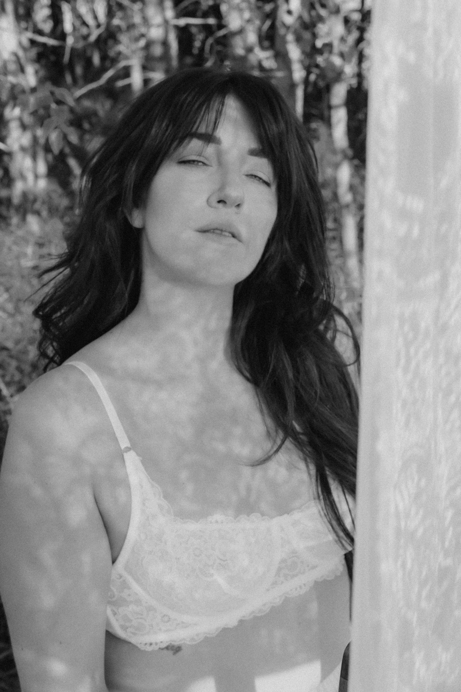 Black and white portrait of woman in lace bra leaning against wall.